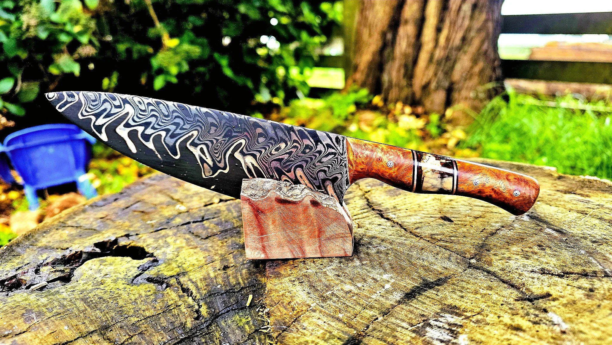A decorative chef's knife with a patterned blade and a wooden handle, resting on a piece of wood outdoors.
