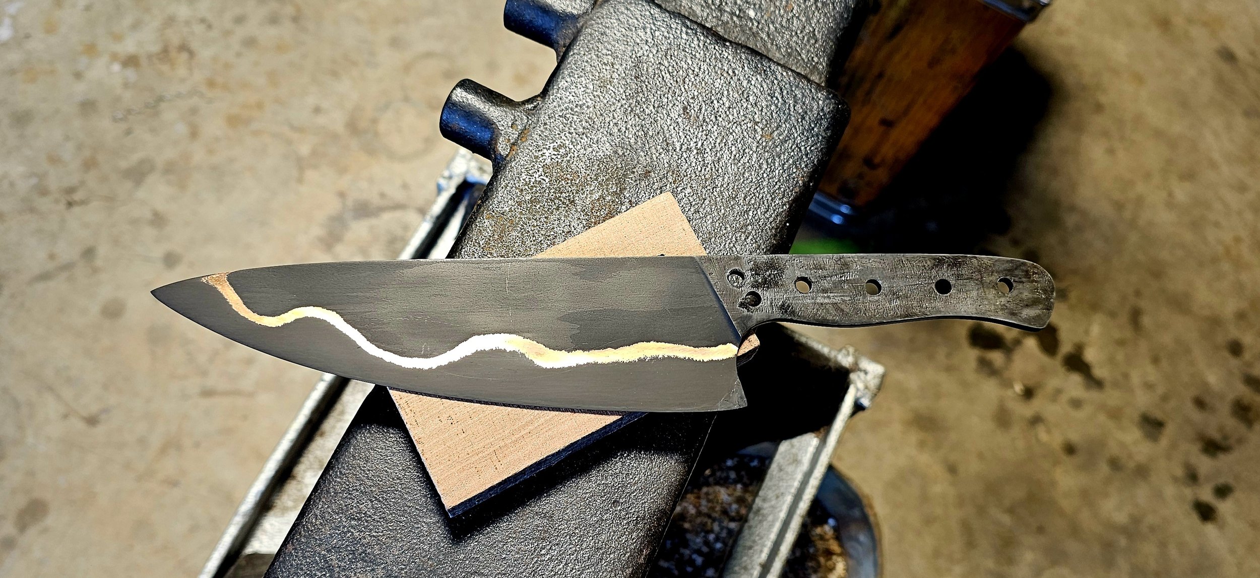 A knifemaking process with a partially finished knife held above a forge, displaying a wavy, layered pattern on the blade.