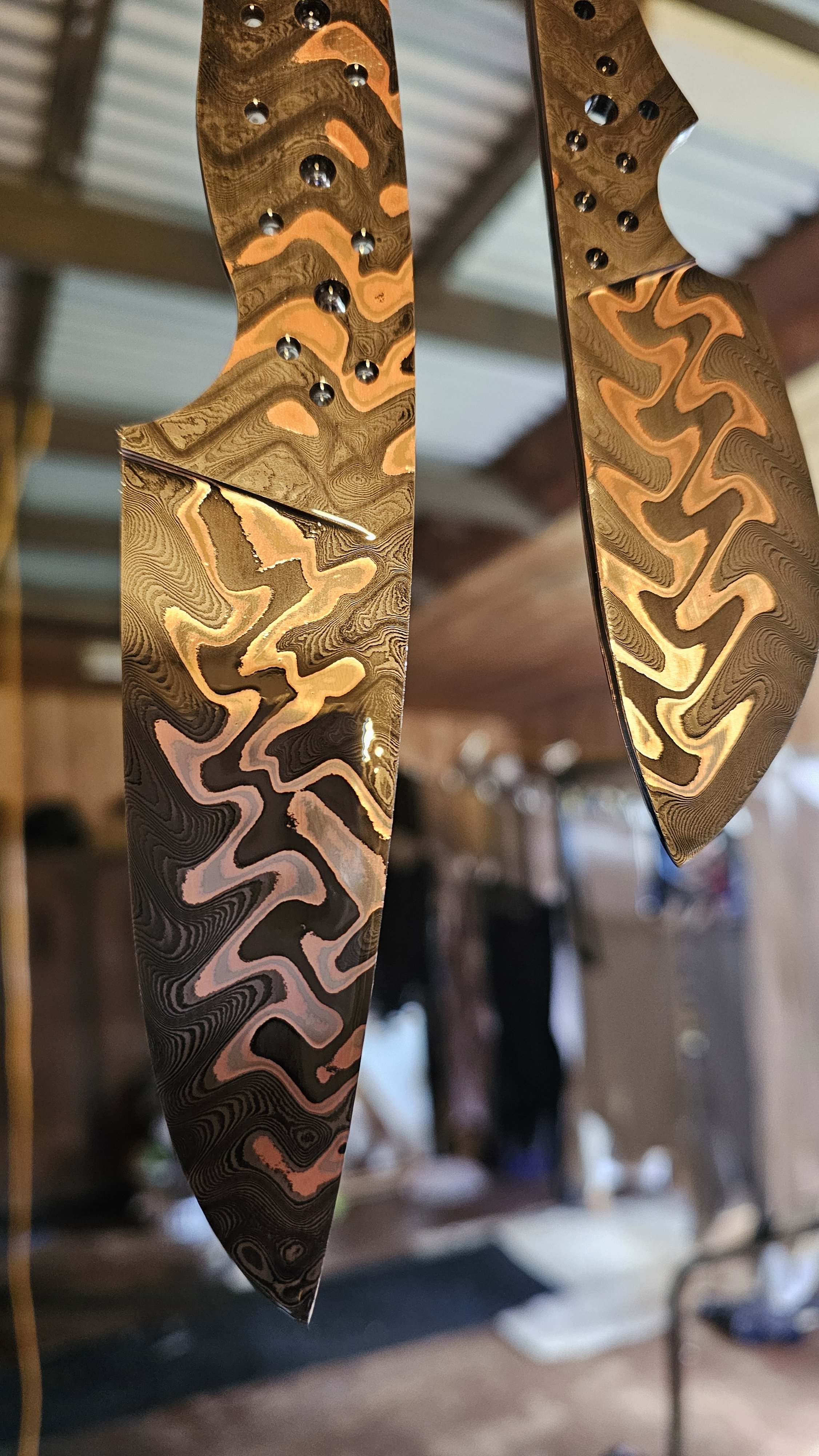 Close-up of two decorative metal blades with swirling, layered patterns and shiny rivets, hanging indoors with a blurred background.