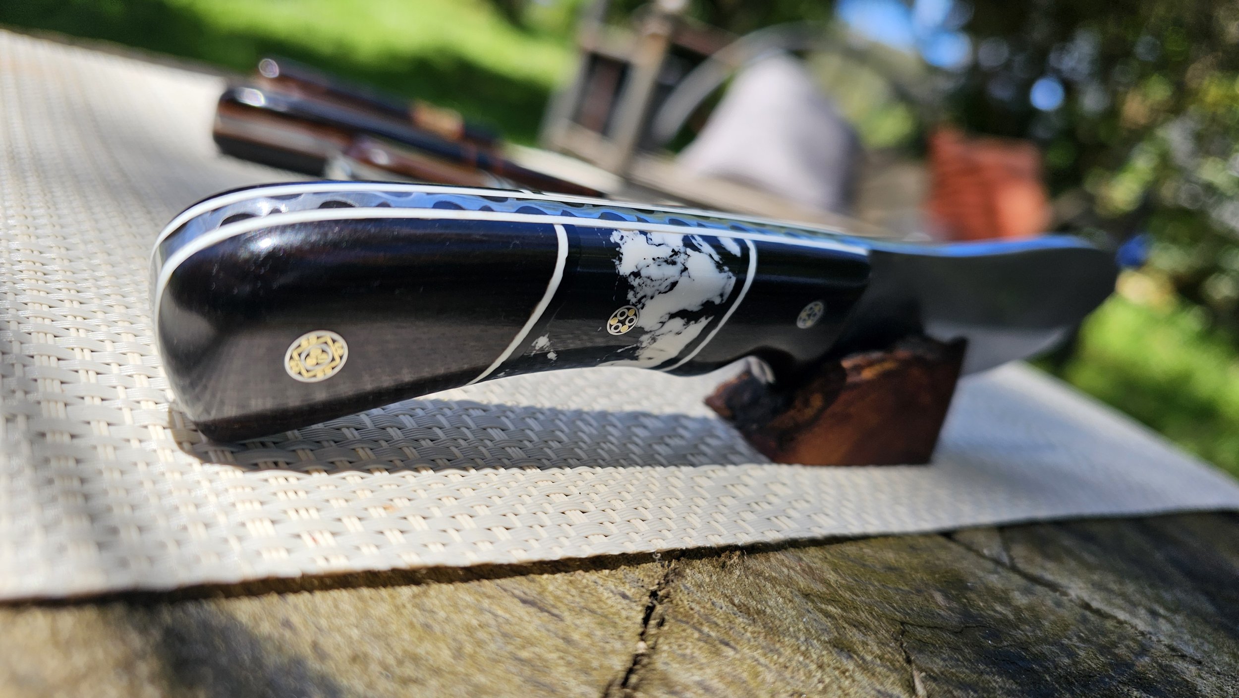 Close-up of a beautiful Nesmuk style knife resting on a white textured surface outdoors. The background is blurred with greenery and some wooden furniture.