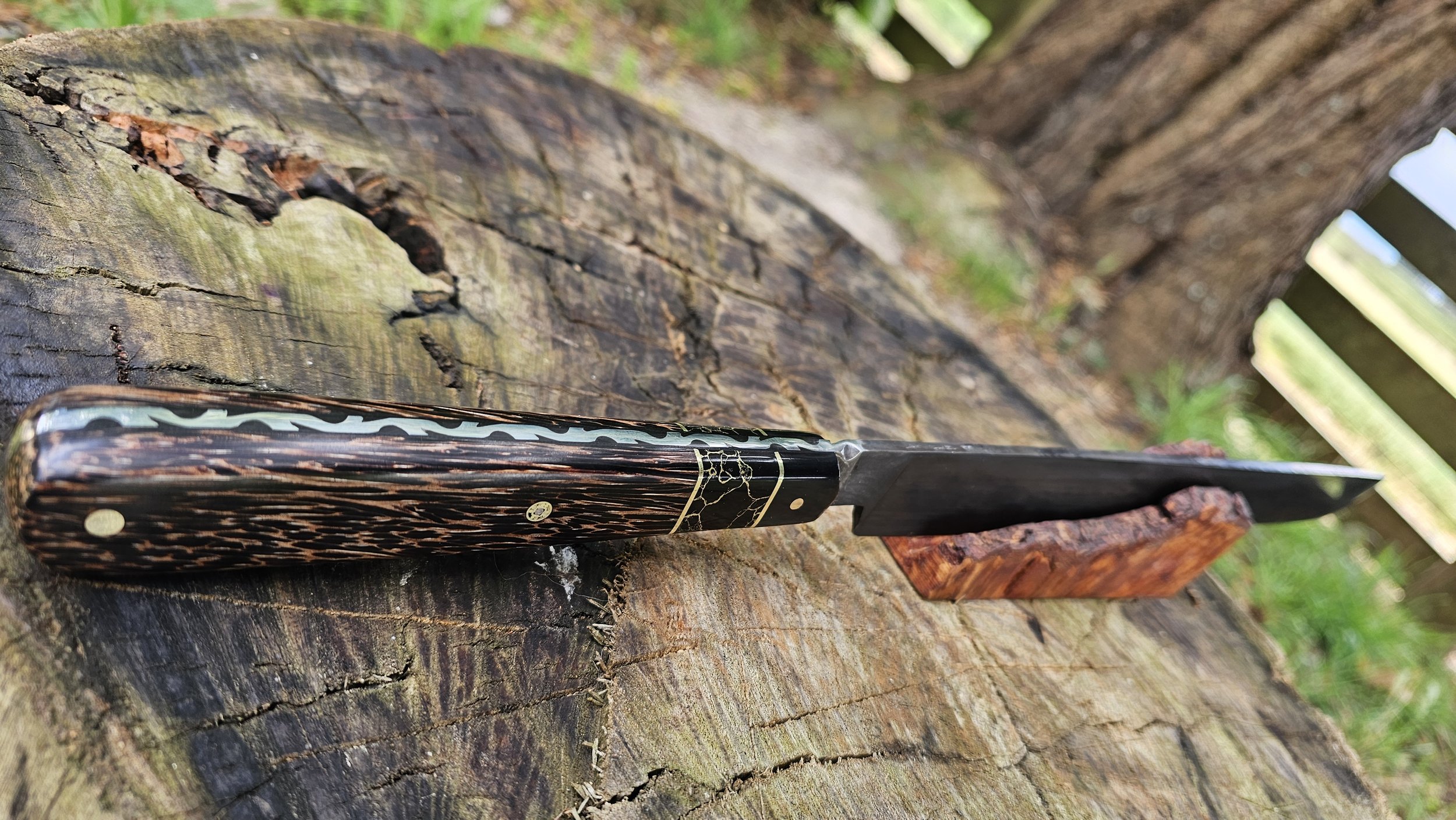 A sushimi sword with a black and speckled handle resting on a weathered wooden log outdoors.