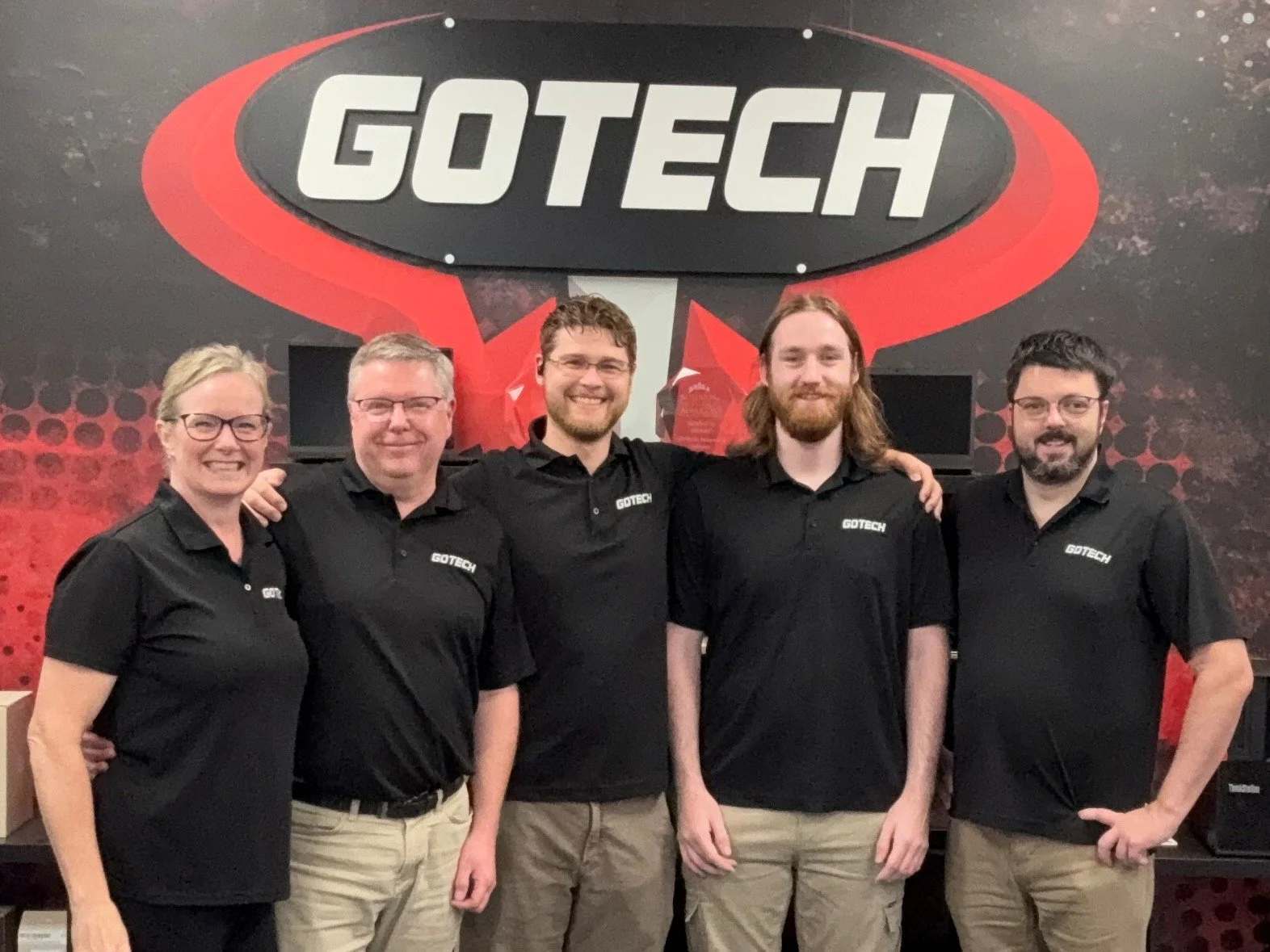 Gotech staff standing in front of a GOTECH sign, all wearing black GOTECH branded polo shirts, smiling, with their arms around each other's shoulders.  Friendly and professional environment.