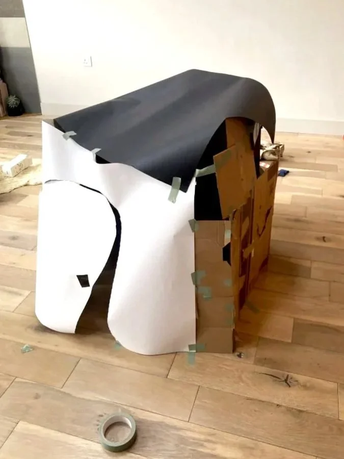 A cardboard den wrapped in black, white, and brown paper, taped together, on a wooden floor in a room with a white wall. Roll of tape on floor.