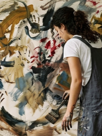 Female artist painting a large abstract canvas with black, brown, red, and blue brushstrokes.