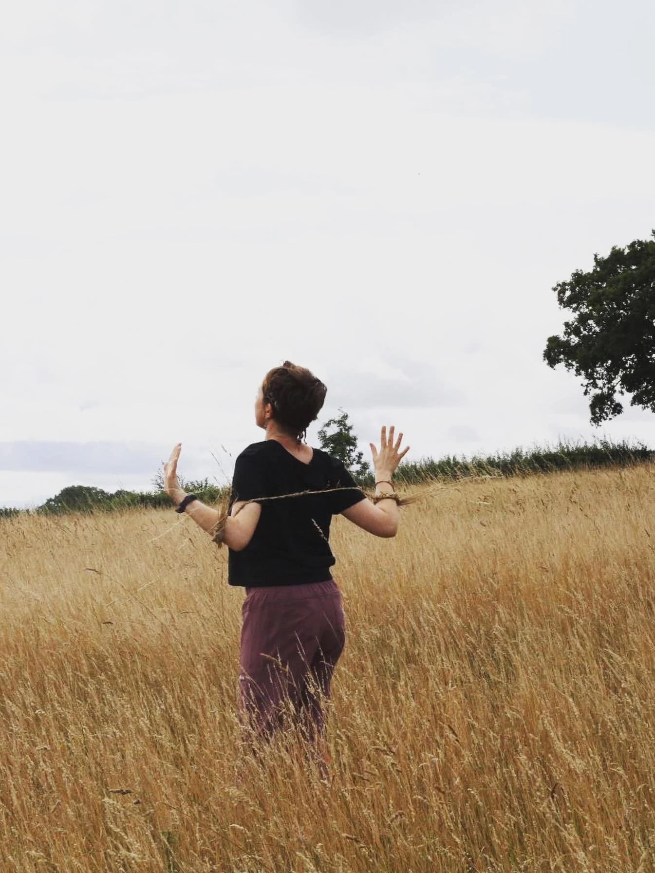 A woman standing in a field of tall, golden grass, with her arms raised, facing away from the camera on a cloudy day.
