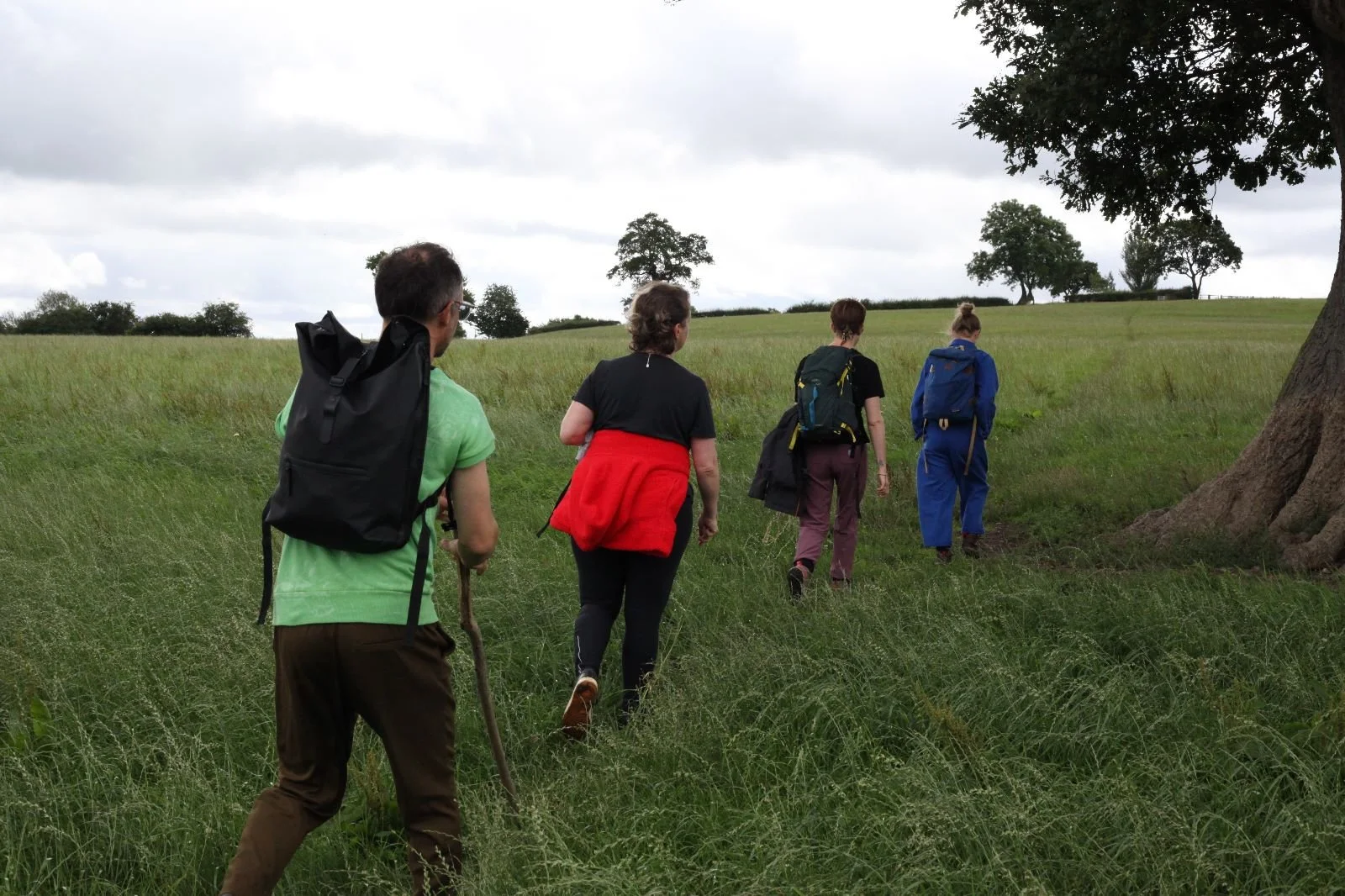 Four people with backpacks walking through a grassy field on a cloudy day, heading toward trees in the distance.