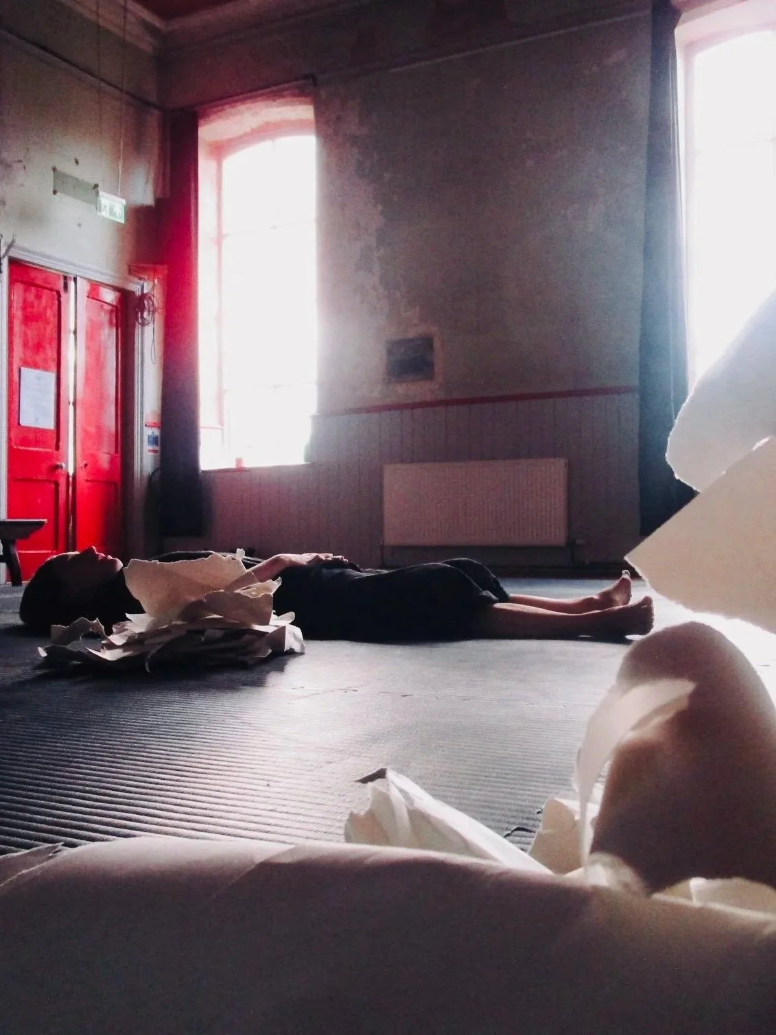 A woman lying on the floor of a room with sunlight streaming through the windows, with crumpled paper scattered around.