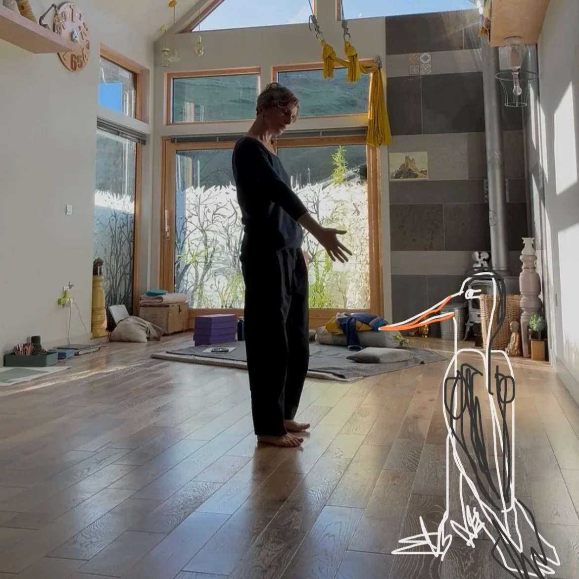 A woman standing barefoot on a wooden floor in a sunlit room with large windows, some yoga mats and cushions on the floor, and a sketch of an umbrella drawn on the floor near her. There are various objects and furniture around, including a clock on the wall and some artwork.