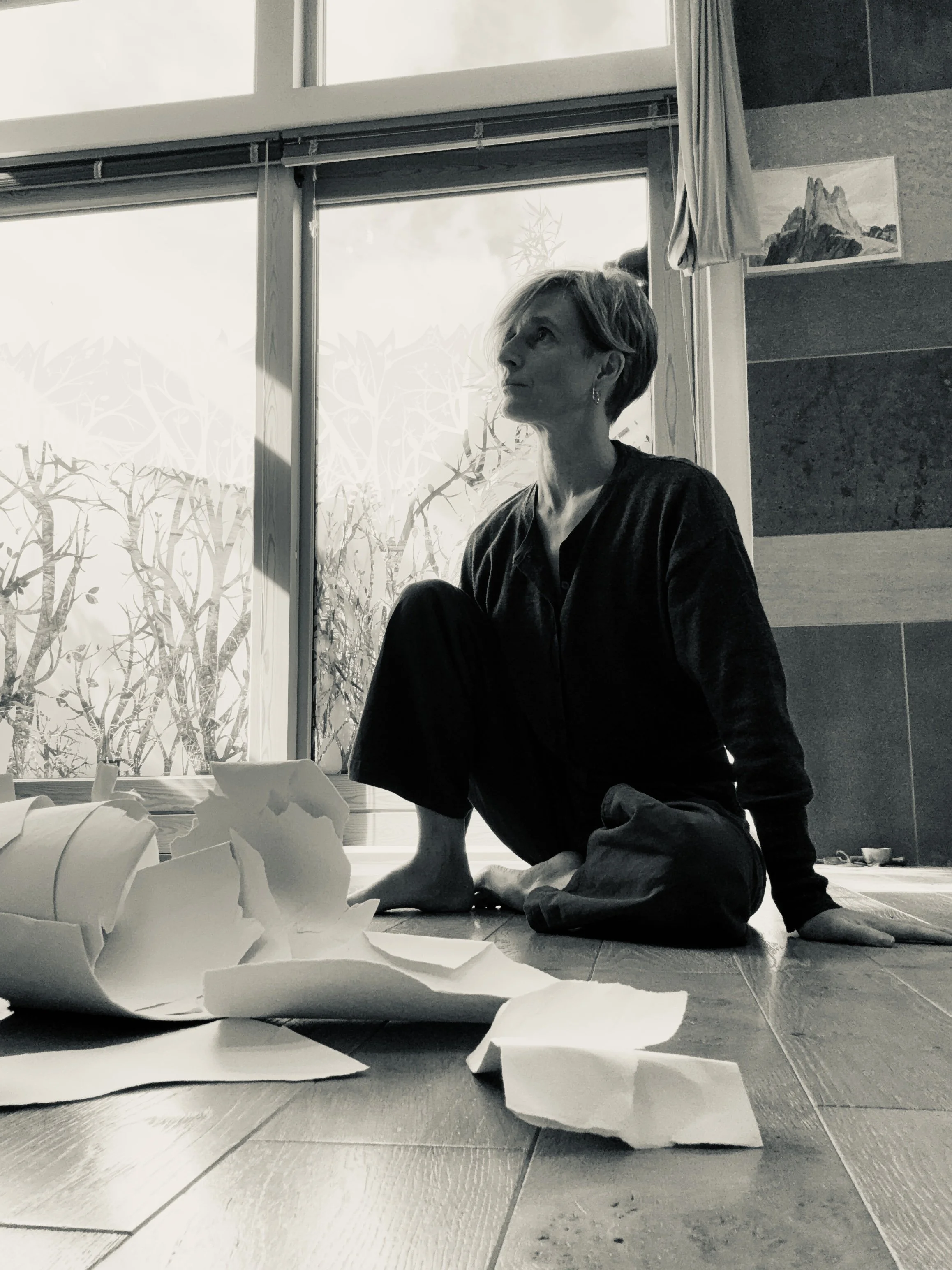 A woman with short hair sitting on the floor next to large sheets of crumpled paper, with a large window and painting of mountains in the background.