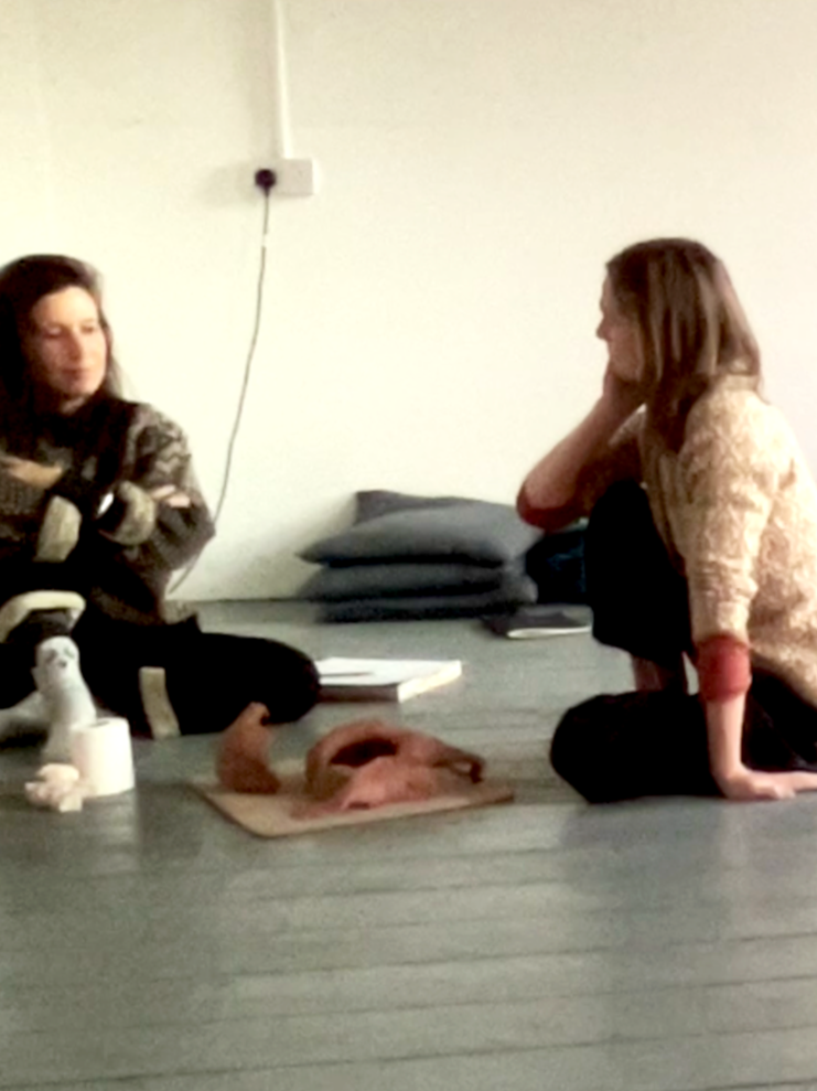 Two women sitting on the floor in a room, facing each other in conversation with clay creation and art materials nearby.