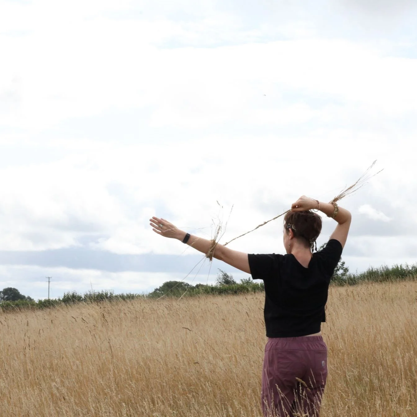 Person standing in a field of tall grass, facing away, with arms extended and holding a stick across shoulders, under a cloudy sky.