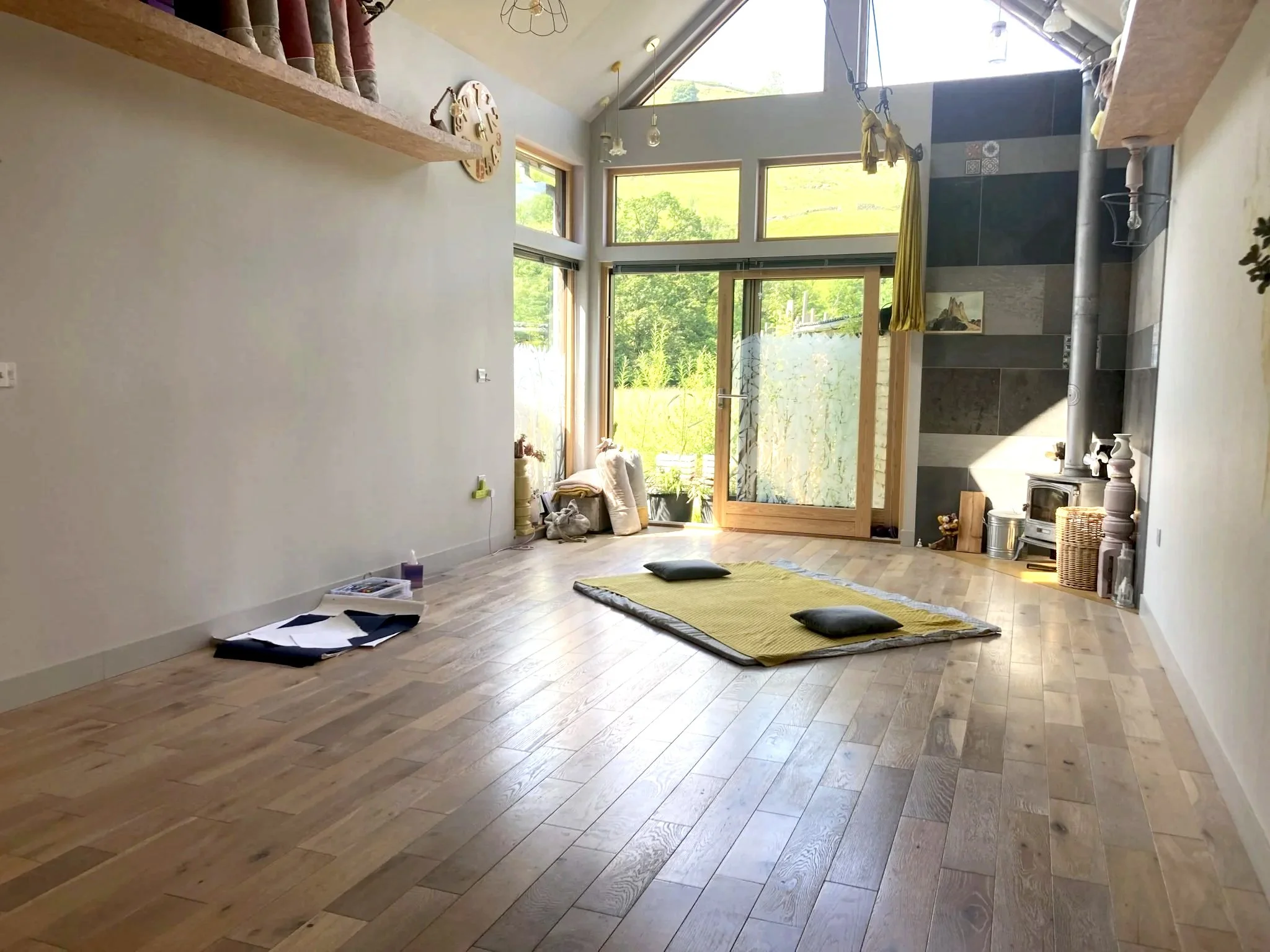Sunny studio with wood flooring, large windows, and a mountain view outside. A soft area for lying down with blankets and cushions for comfort. Paper and art materials on the floor, indicating creative process.