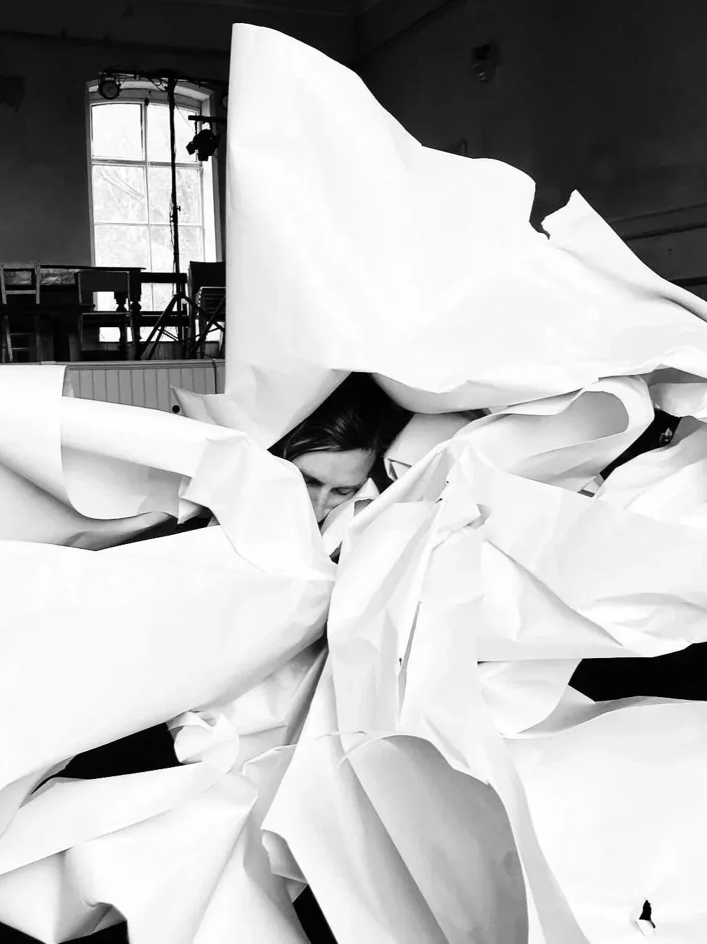 Black and white photo of a person surrounded by crumpled paper, with a large window and studio equipment visible in the background.