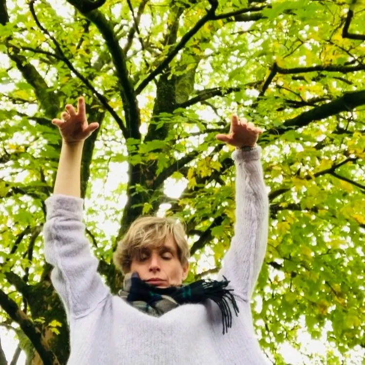 A person with blonde hair wearing a light sweater and a plaid scarf, standing outdoors beneath a tree with green and yellow leaves, with arms raised.