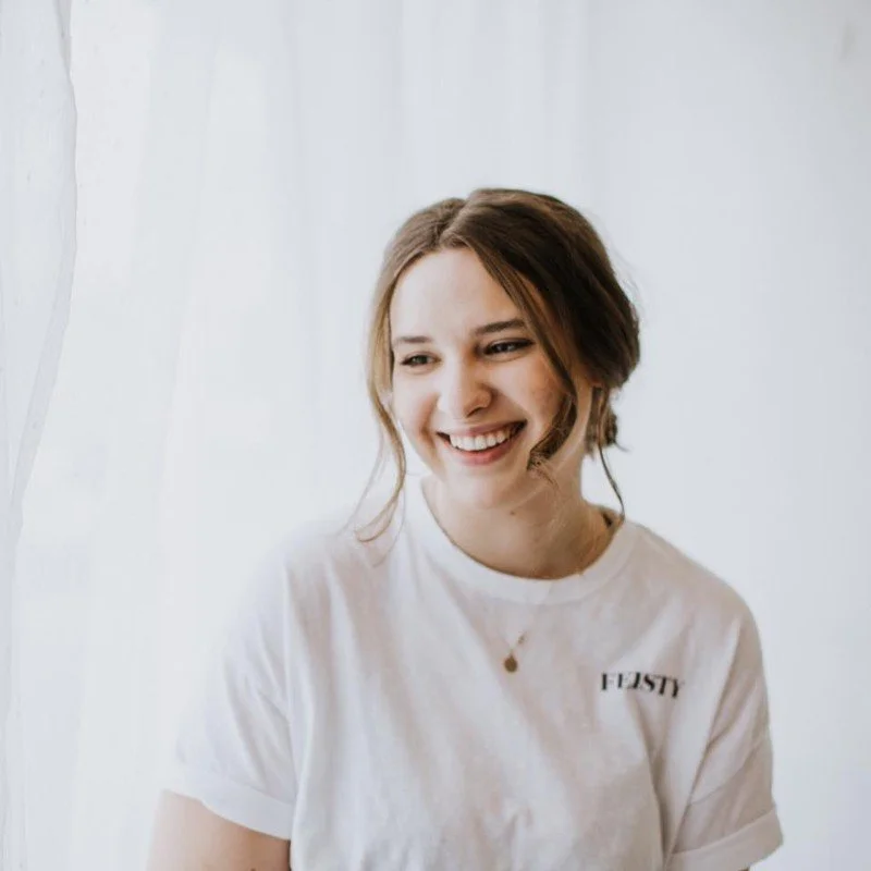 A young woman with shoulder-length brown hair is smiling and looking to her right. She is wearing a white t-shirt with a small text on the chest and a necklace with a small pendant. The background is bright and plain.