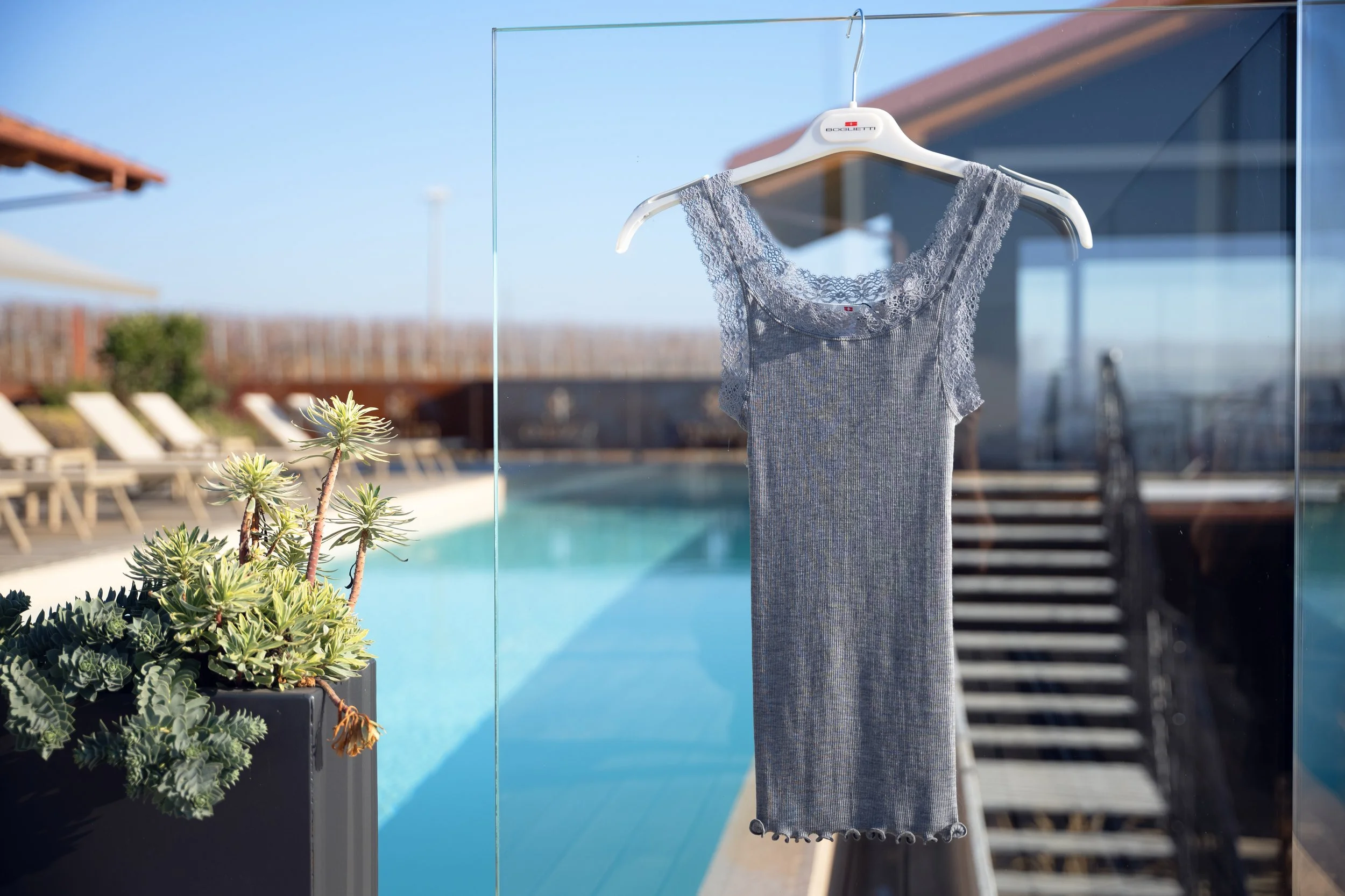 A lace and gray dress hanging on a white hanger outside on a glass wall with a swimming pool and lounge chairs in the background.