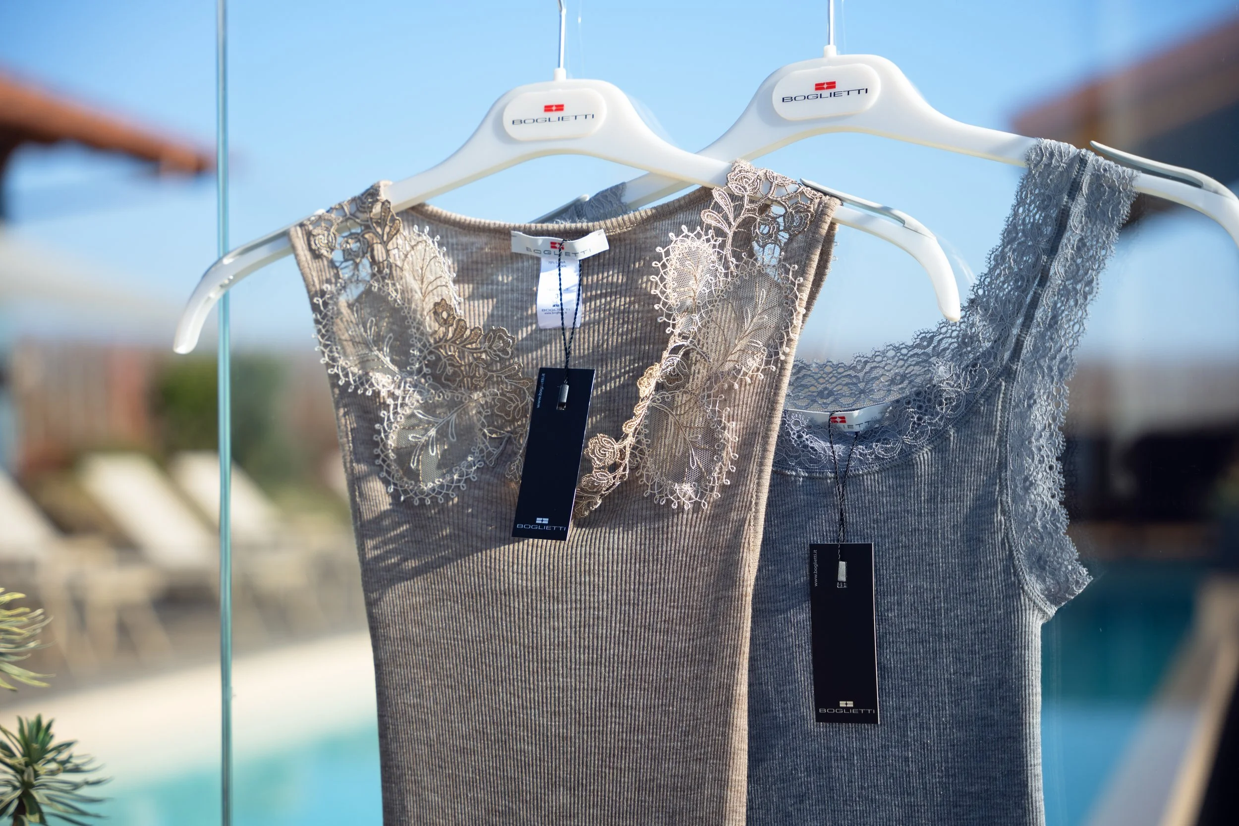 Lace and embroidered tops hanging on white BOGLIETTI brand hangers outdoors.