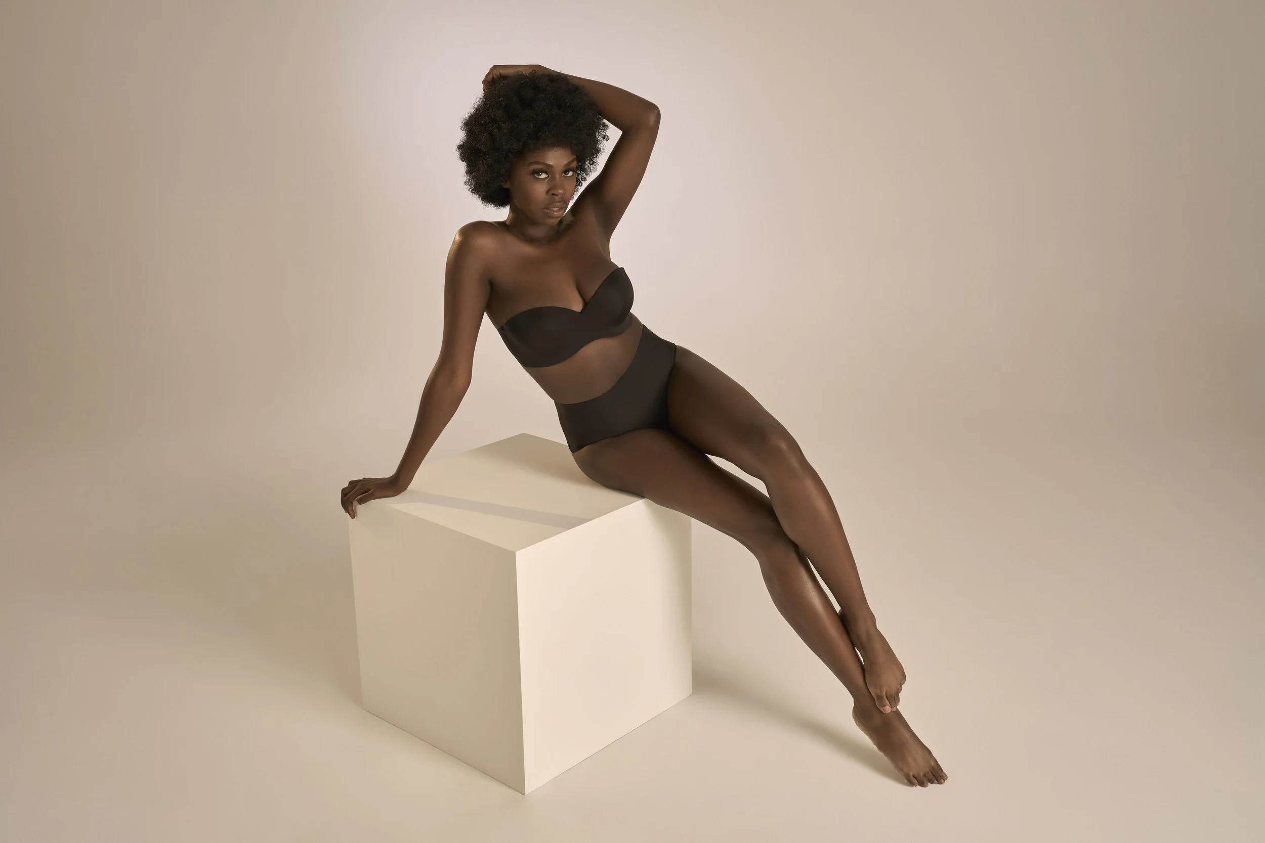 A woman with dark skin and curly hair in black lingerie, posing sitting on a white block against a plain beige background.