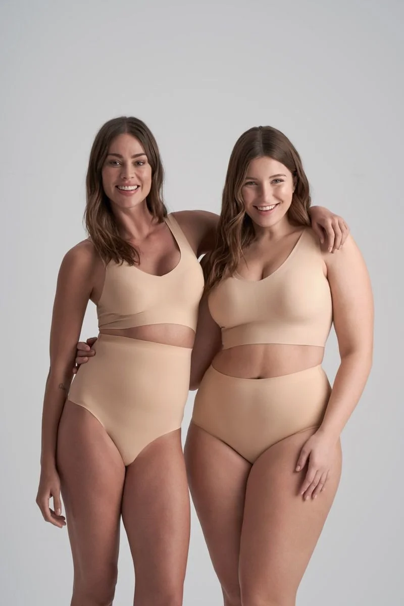 Two women standing close together, smiling, wearing matching beige underwear, with one woman’s arm around the other’s shoulder, against a plain light gray background.