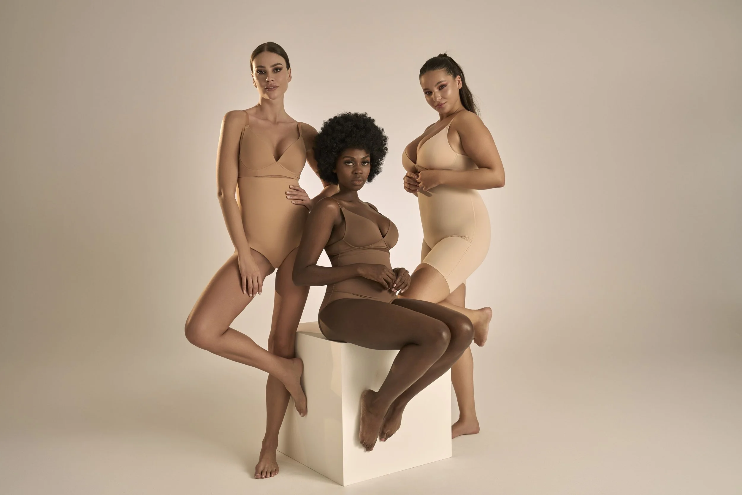 Four women of diverse ethnicities with different skin tones pose together in nude-colored shapewear against a neutral background. Two women are standing, one is seated on a white box, and one is sitting on the ground with her leg raised. They look co
