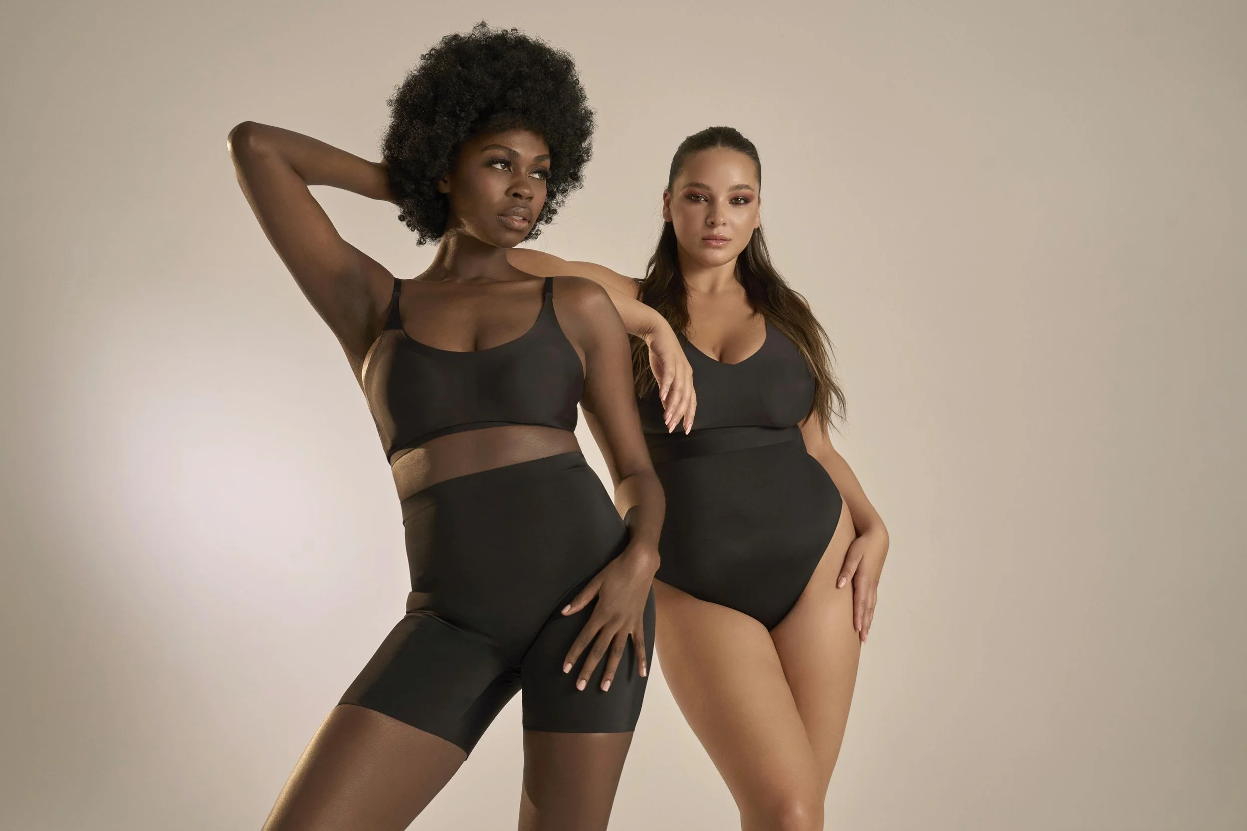 Two women wearing black lingerie pose against a plain light-colored background.
