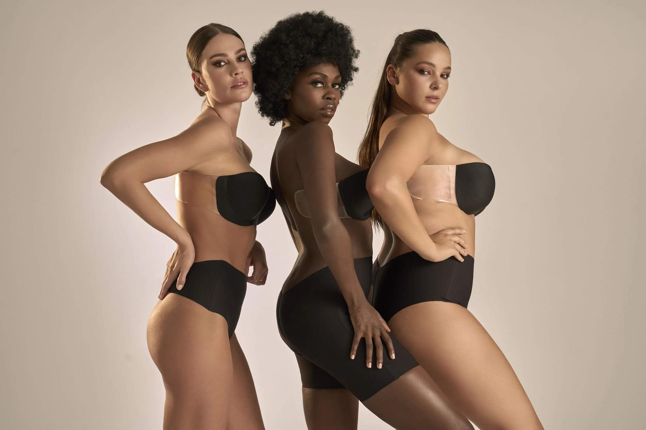 Three women with different skin tones, wearing black and beige strapless top and shorts, standing in a line against a plain background, facing slightly to the side with one arm on their hip.