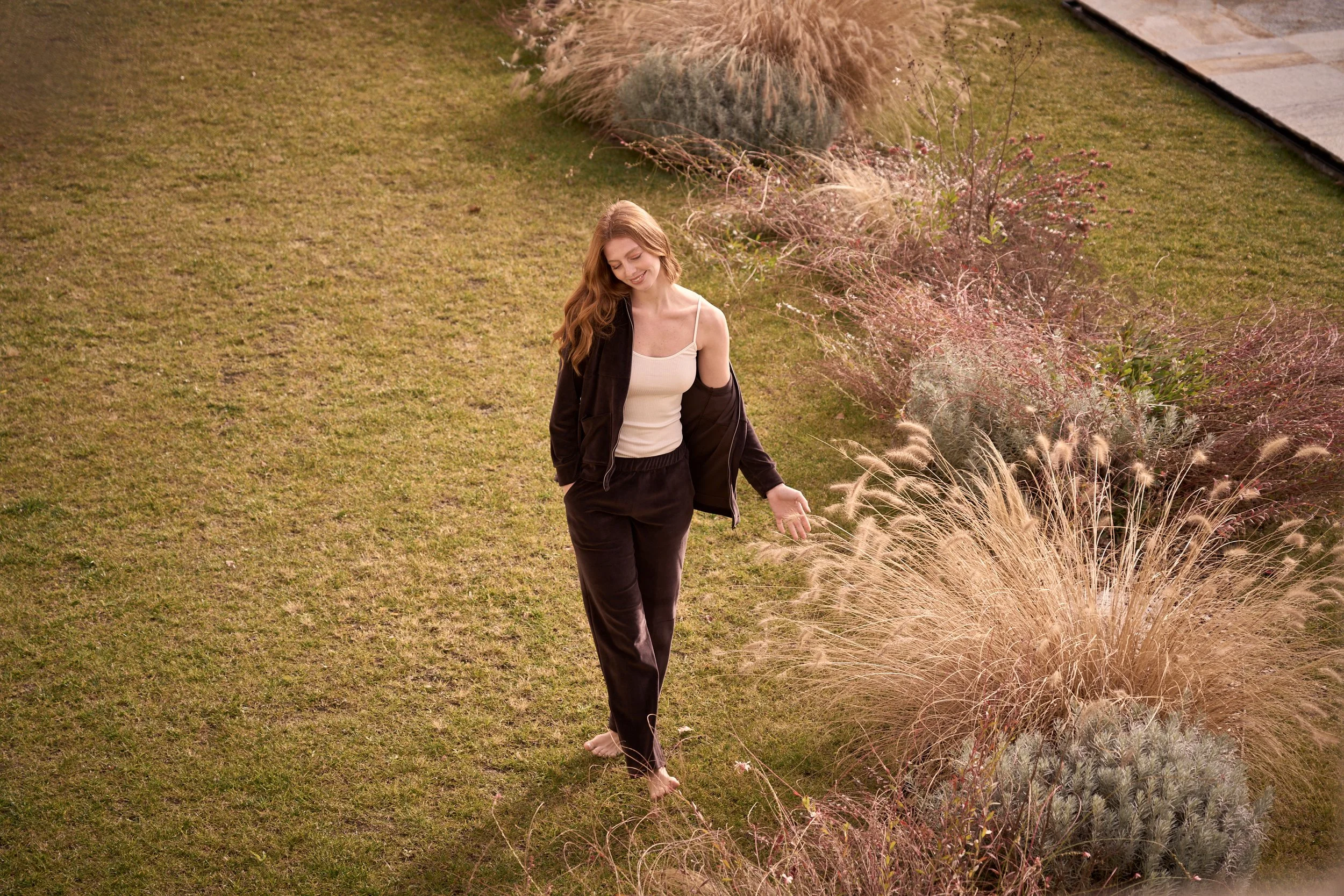 A woman with long red hair walking barefoot on a grassy lawn, smiling with her eyes closed, wearing a cream tank top and black pants, with a dark jacket draped over her shoulder. She is surrounded by ornamental grasses and plants.