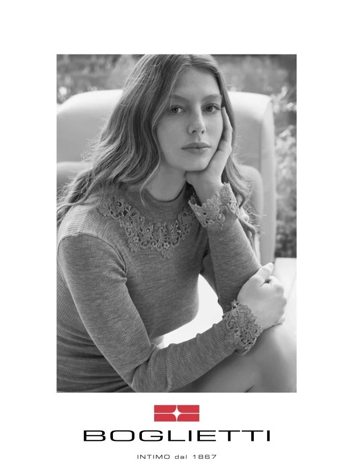 A black and white portrait of a young woman with long wavy hair, resting her head on her hand, wearing a sweater with lace details, sitting outdoors with a blurred background. Below the photo is a logo and the text "BOGLIETTI" and "INTIMO dal 1867".