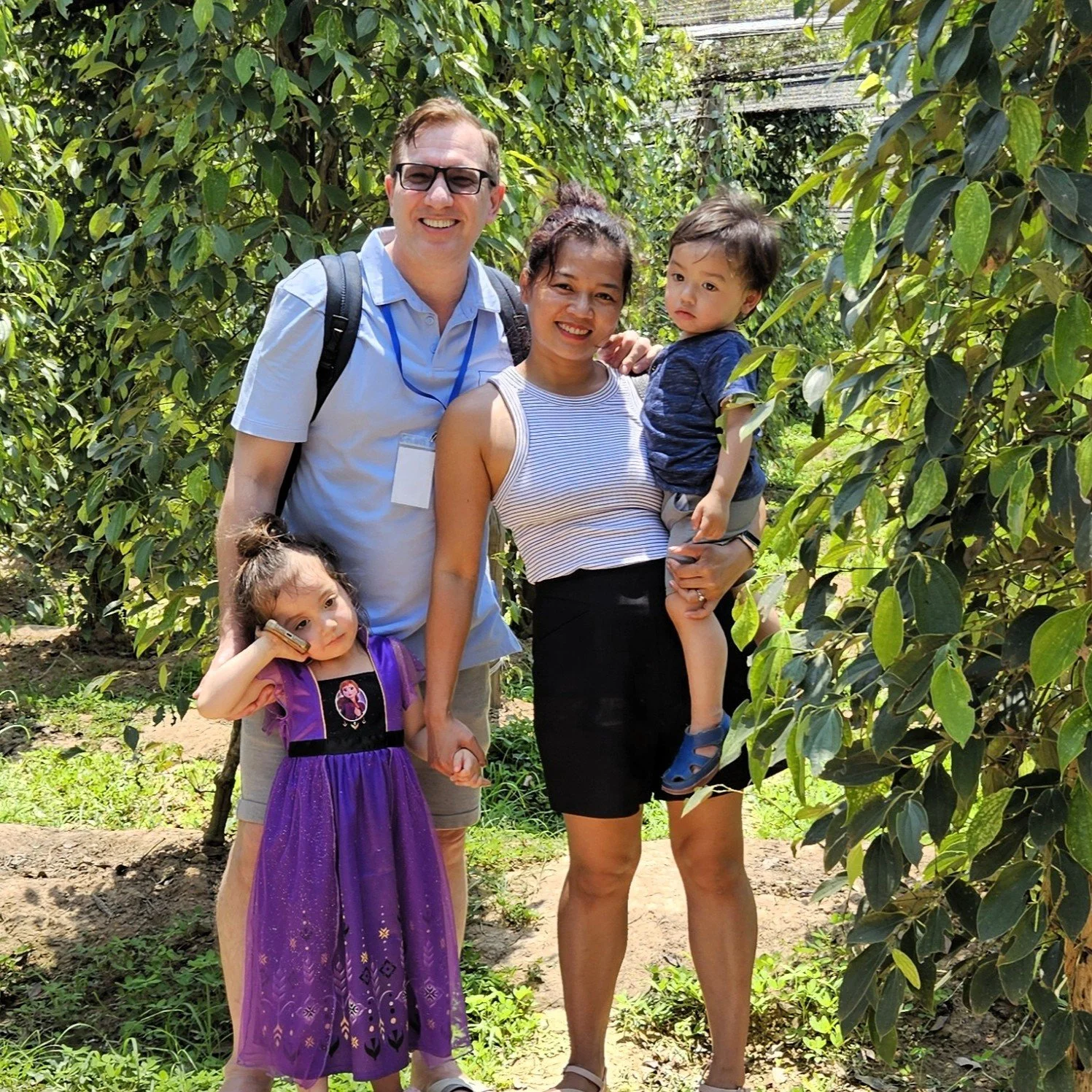 A group of four people, including one adult woman, one man, and two young children, standing together in a lush green orchard or farm with rows of leafy trees, smiling and posing for the photo.