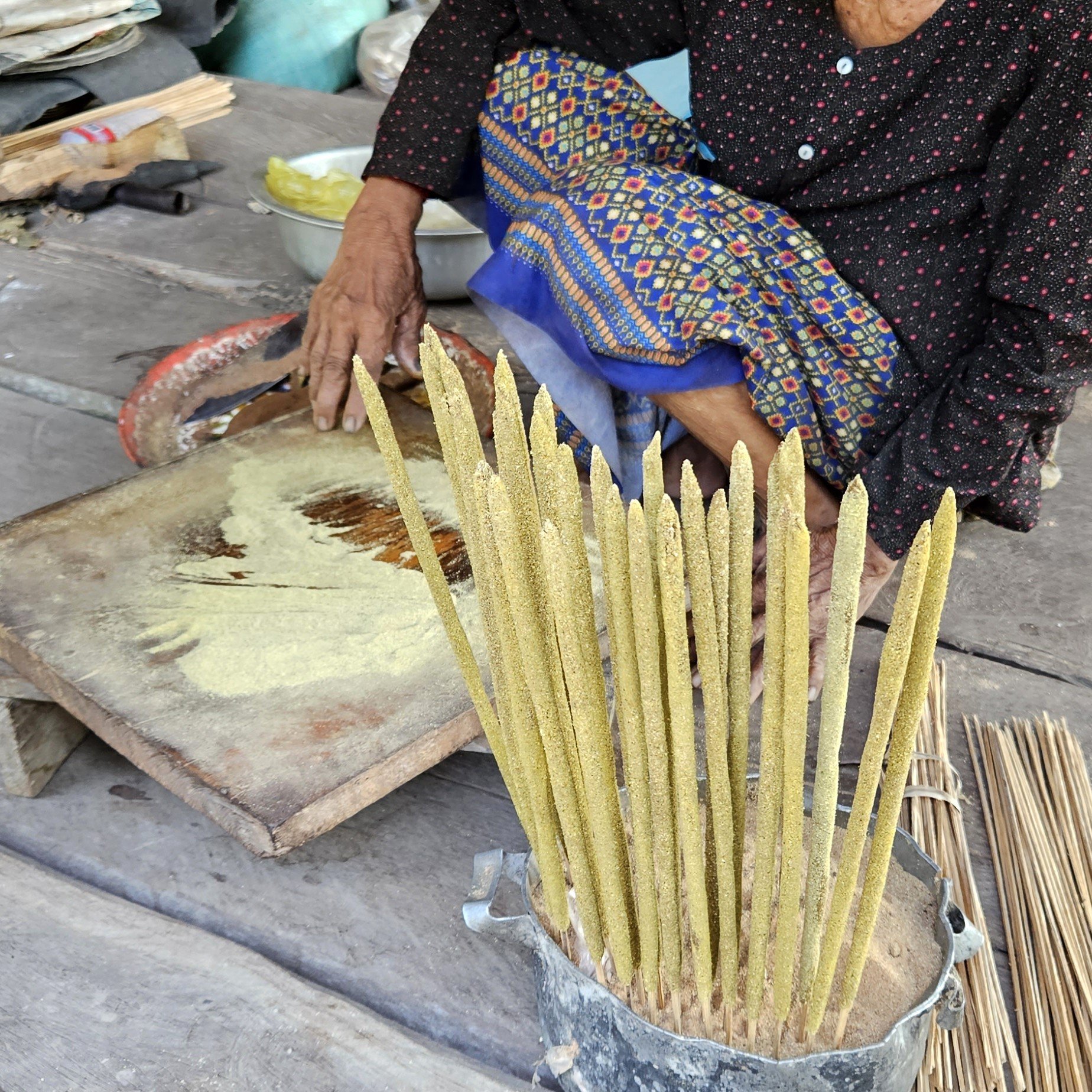 An elderly person wearing a black top with red pattern and colorful patterned pants sits at a rustic wooden table, making traditional food using a round griddle, strips of sticky rice, and banana leaves.