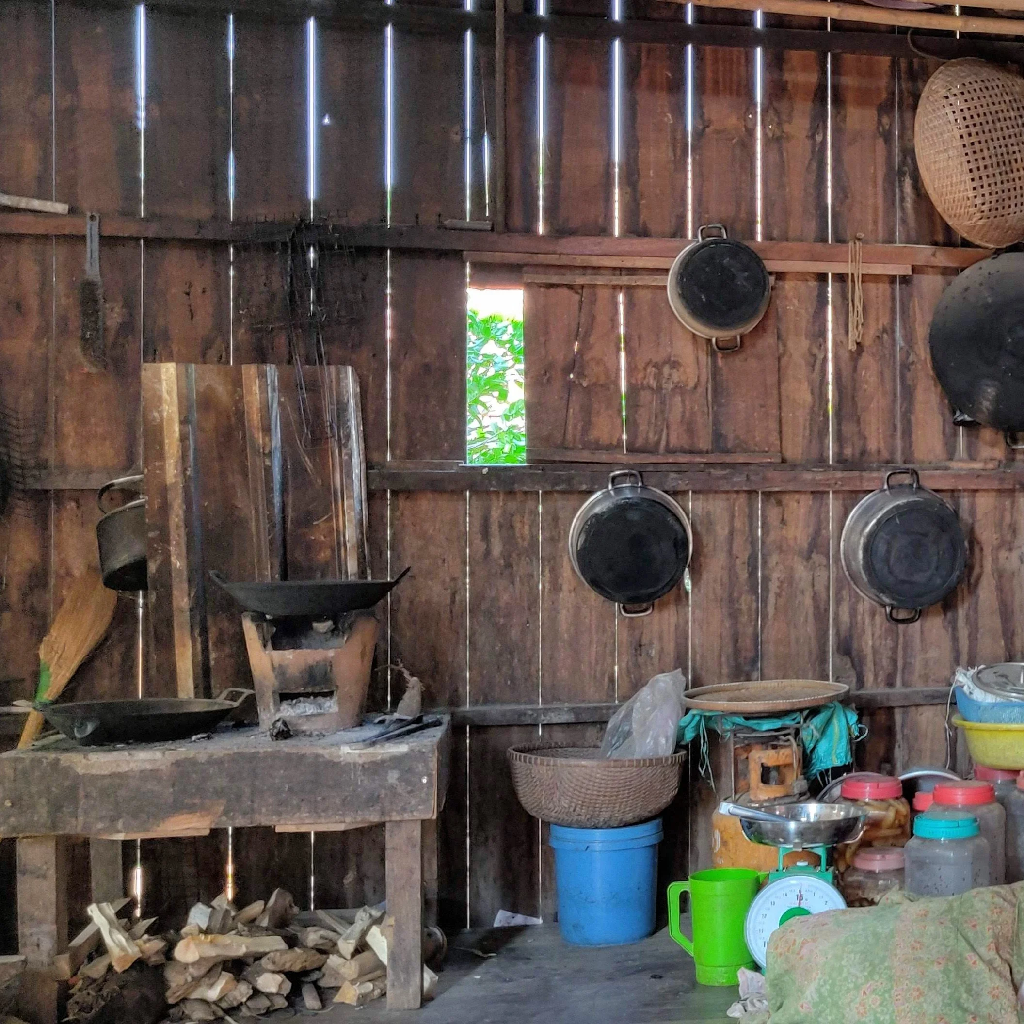 A wooden shed interior with stacked firewood, various buckets, containers, and kitchenware. A small window allows natural light inside.