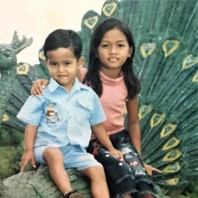 A young boy and girl sitting on a large green plant or tree in front of a background with more greenery. The boy is wearing a light blue uniform with a patch and the girl is wearing a pink top and black skirt with a pattern.