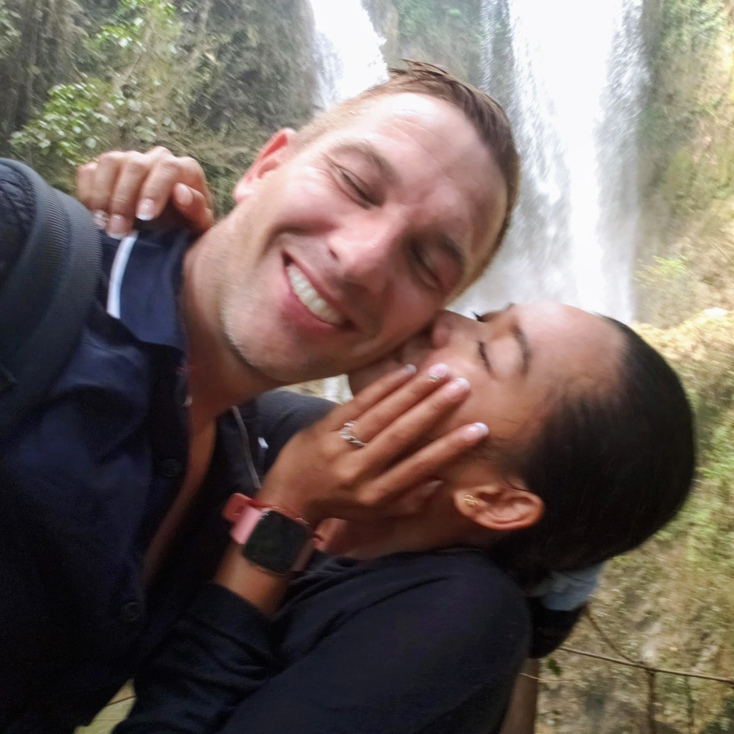 A smiling man with glasses and short brown hair taking a selfie outdoors near a waterfall, with greenery around.