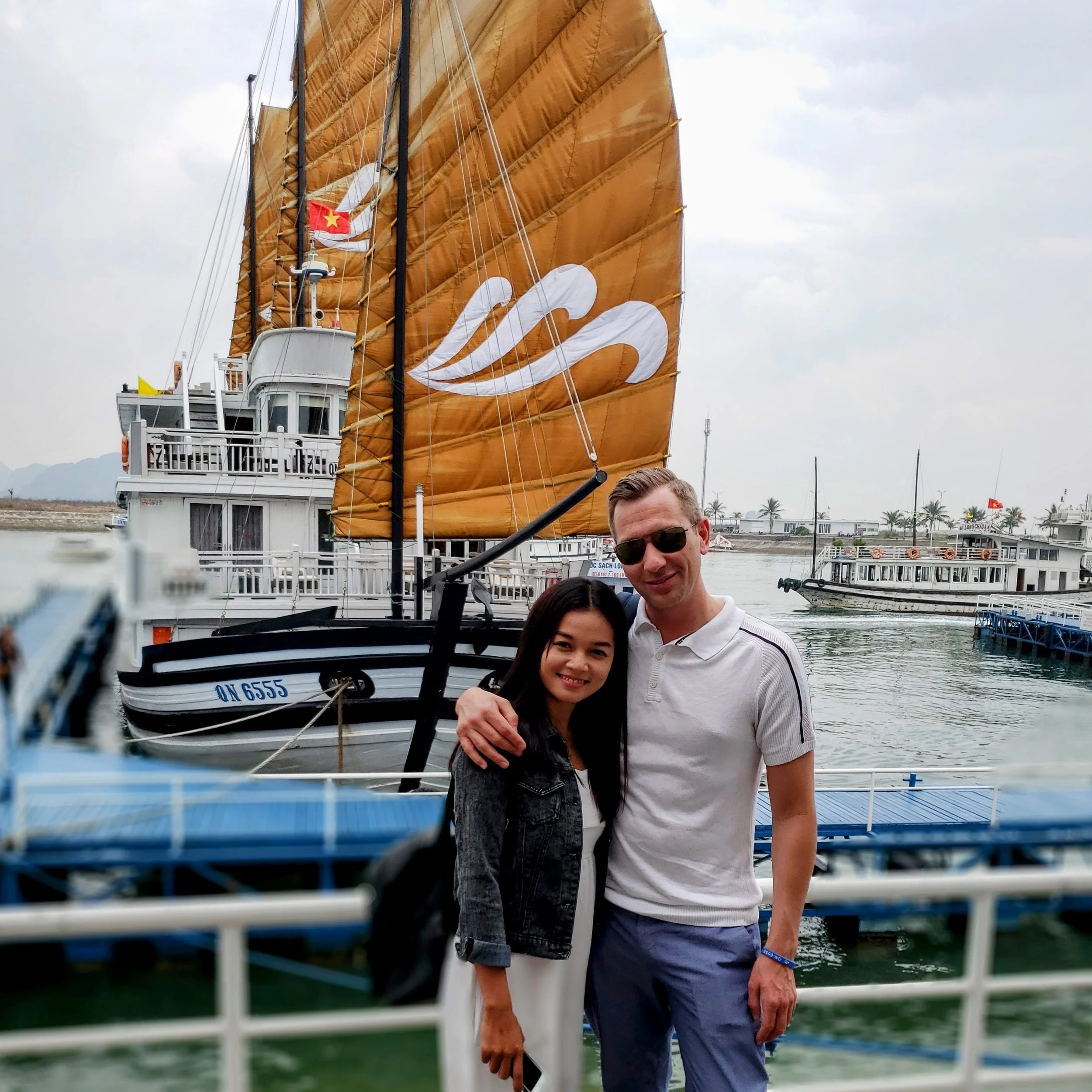 A group of people on a boat dock, with a boat featuring a large orange sail and a red flag with a yellow star in the background.