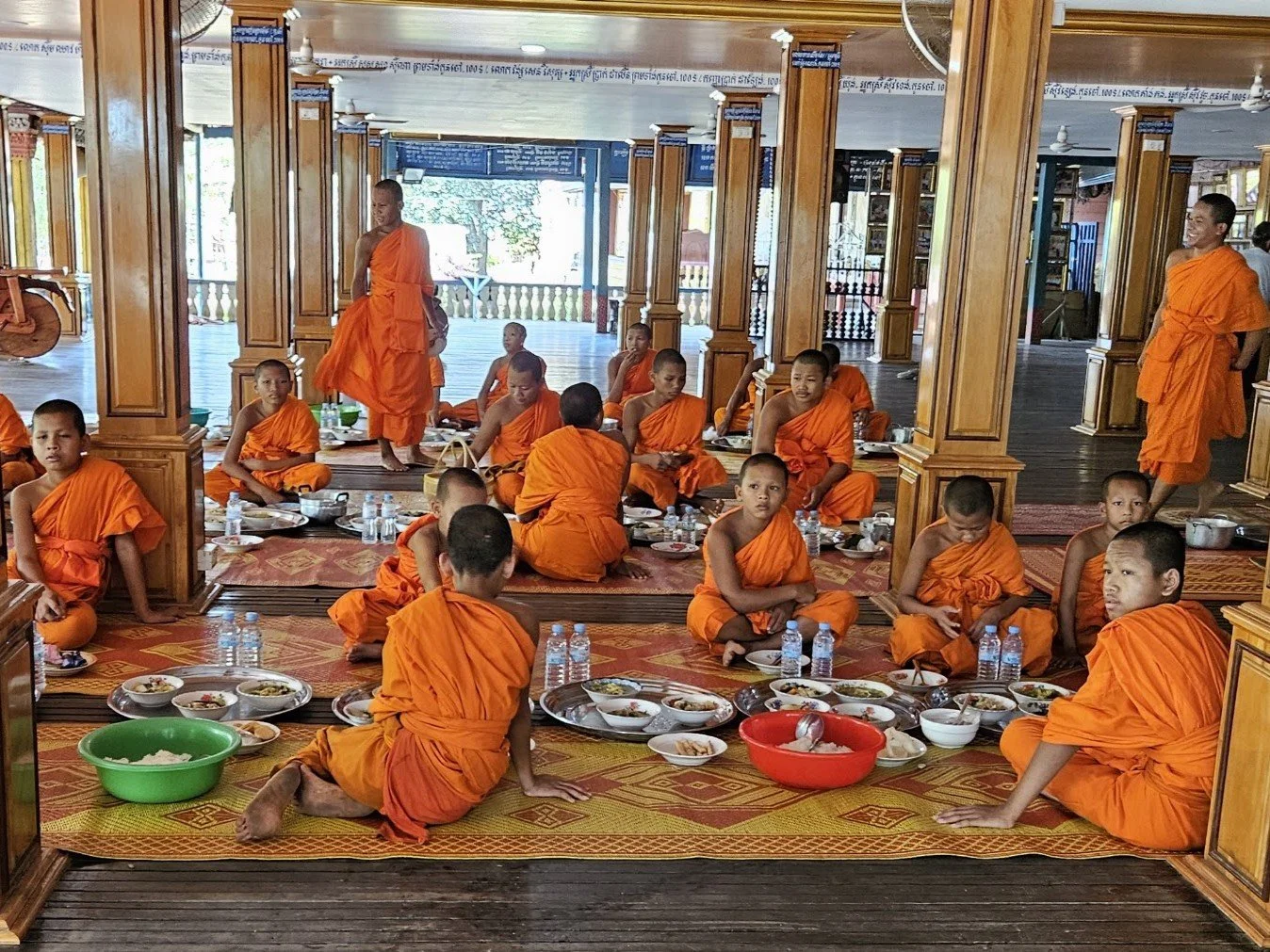 Young Buddhist monks in orange robes sitting on a carpeted floor inside a wooden temple, with some standing or walking, and bowls of food in front of them.