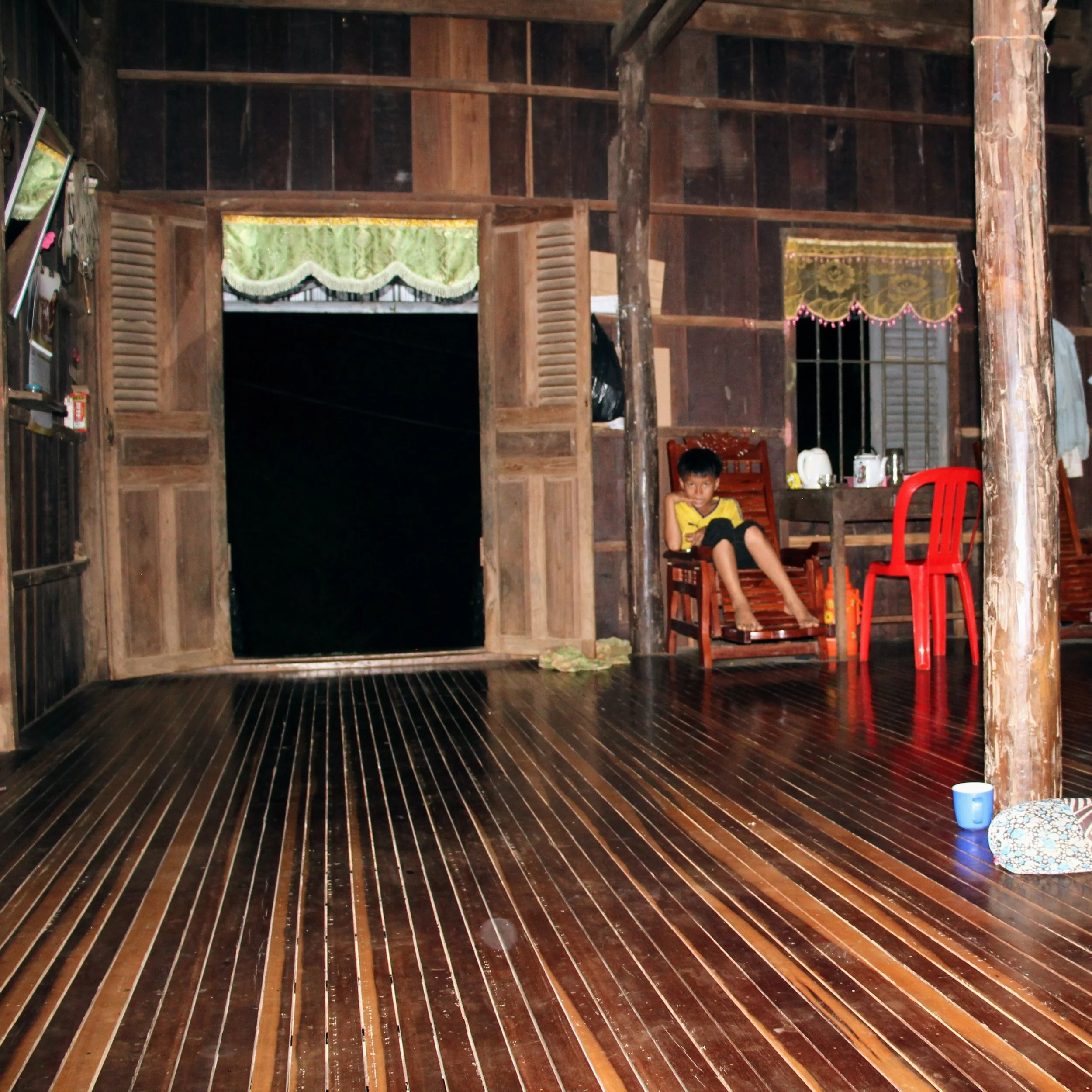 Interior of a wooden house with dark polished wooden floors, walls, and ceiling, green and yellow curtains, a clear window, colorful chairs, a boy sitting on a wooden bench, and various household items.