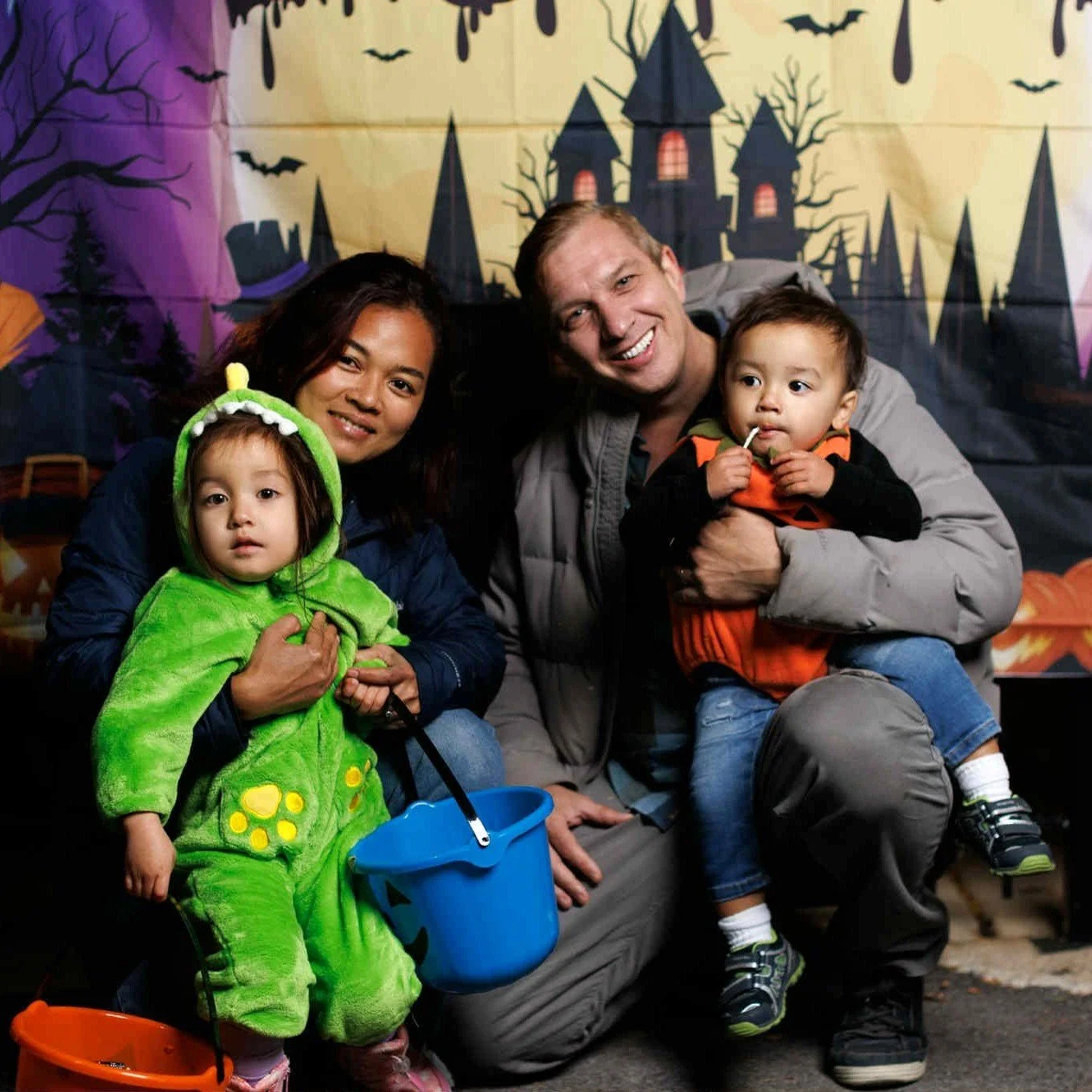 A family dressed in Halloween costumes, with a child in a green dinosaur costume, an adult woman, an adult man, and a young child dressed as a pumpkin, in front of a Halloween-themed background with haunted house, bats, and trees.