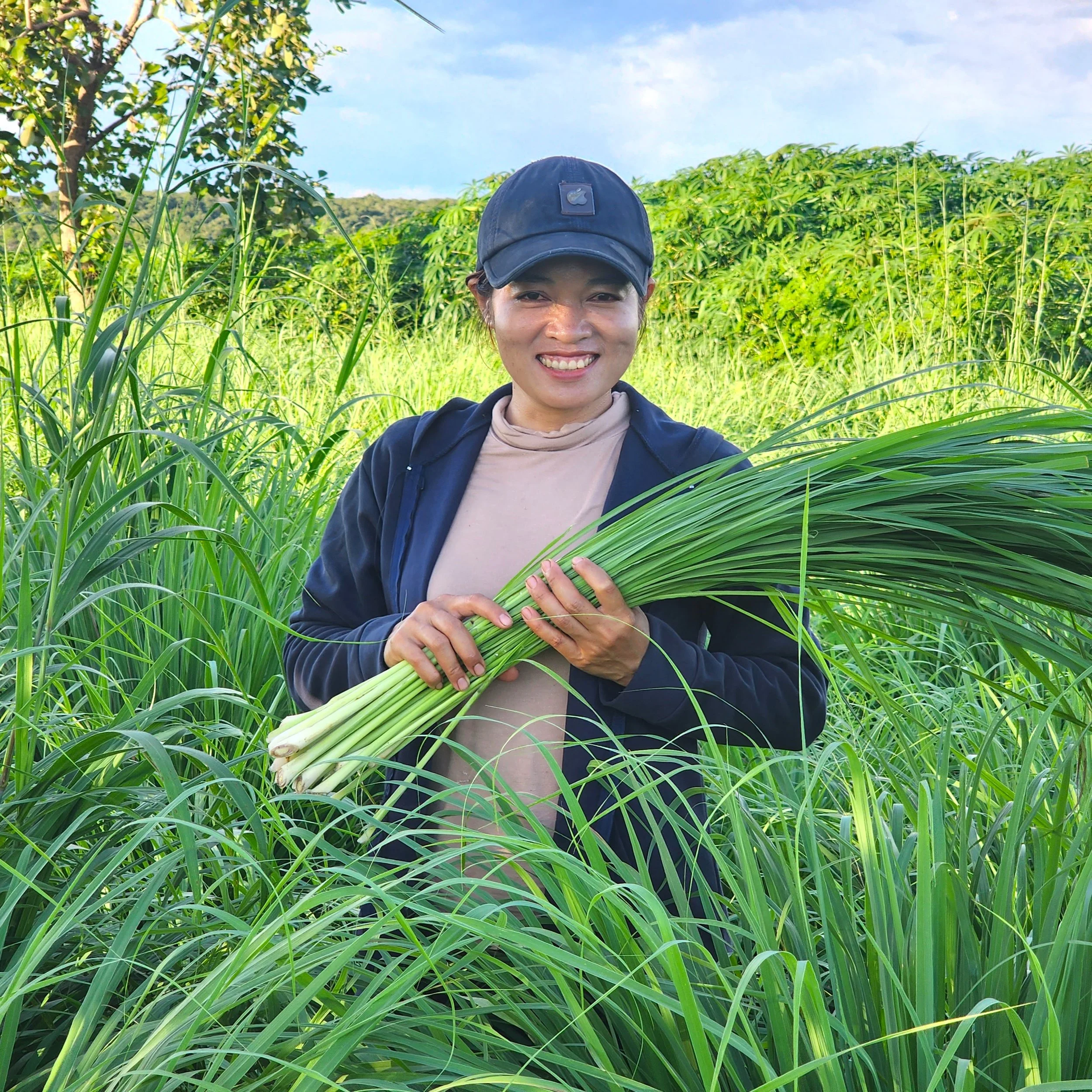 A woman smiling outdoors surrounded by tall green lemongrass and plants, with trees and a blue sky in the background.