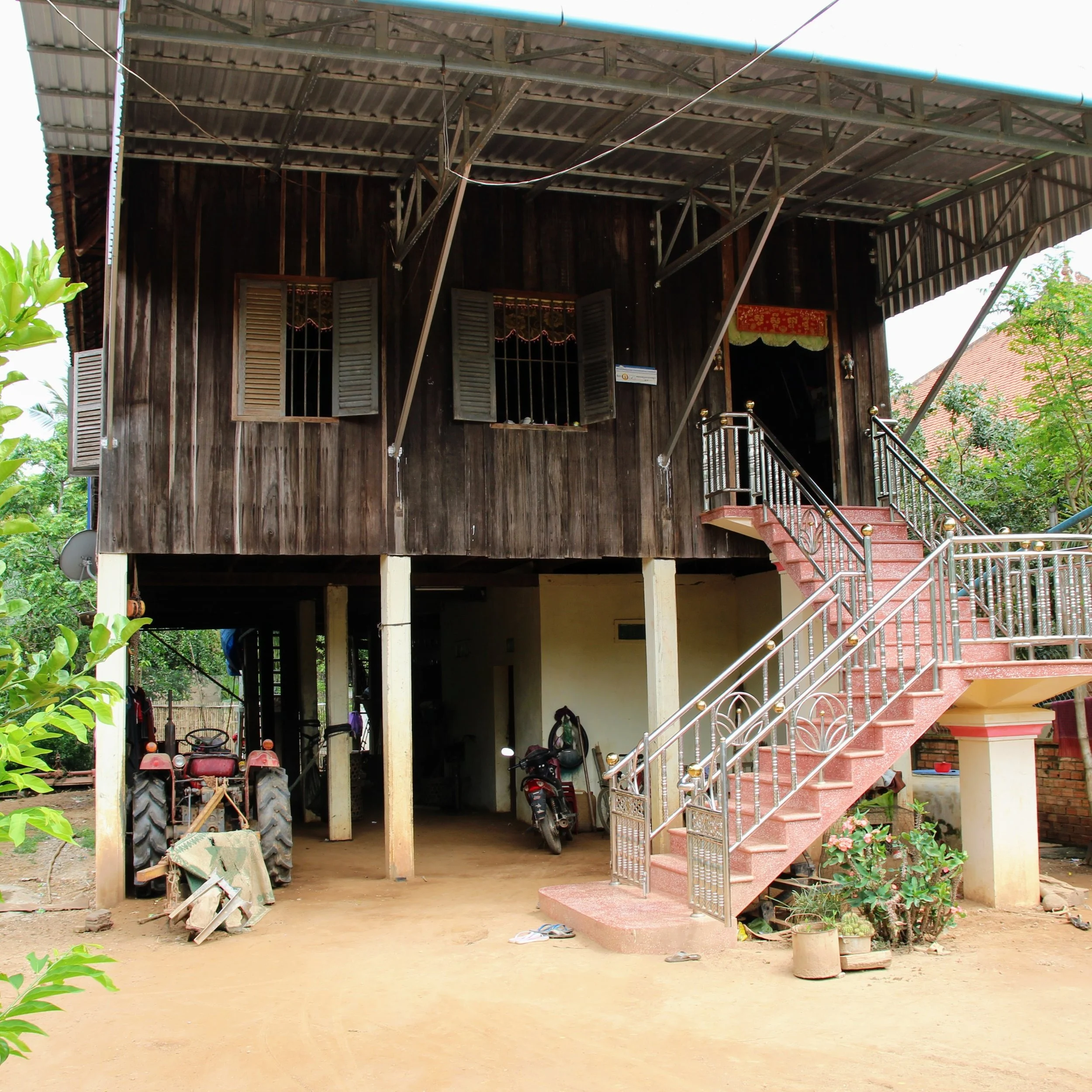 The image shows a two-story house with a wooden exterior, open windows with shutters, and a staircase leading up to the entrance. The upper floor has a metal roof supported by metal beams, and the lower part is open underneath, used as a storage or parking area for tractors and motorcycles. There is greenery around the house and various objects on the ground.