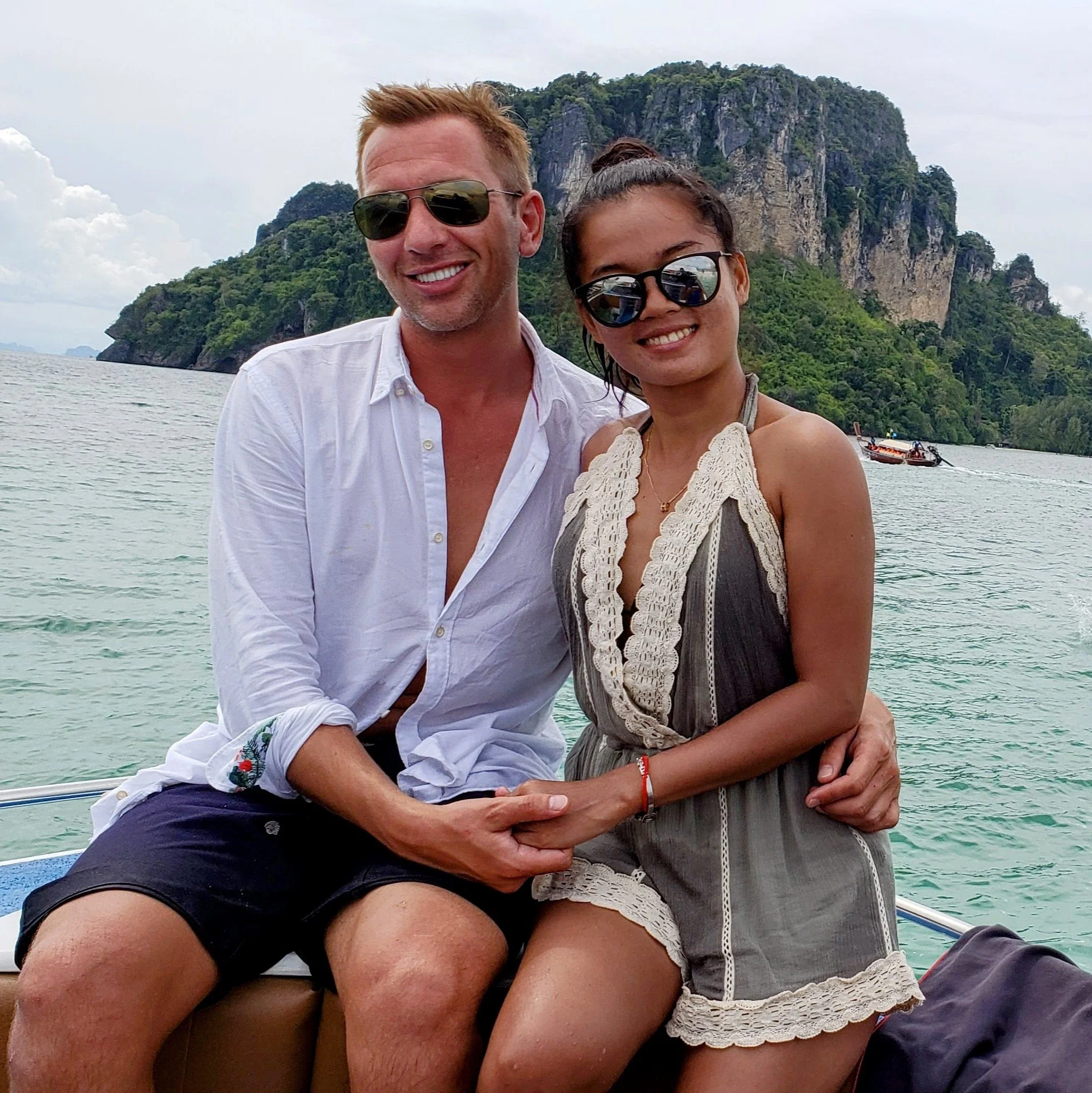 A smiling man with sunglasses and a white shirt, and a smiling woman with sunglasses and a lace dress, sitting on a boat with a scenic island and water in the background.