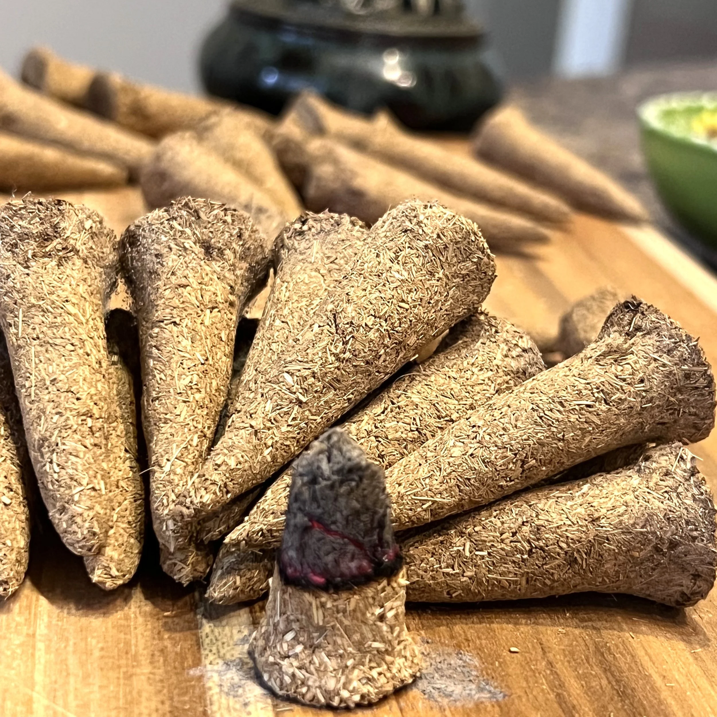 Close-up of a pile of compressed herbal tobacco sticks with a blurred background that includes a green bowl and a dark object on a wooden surface.