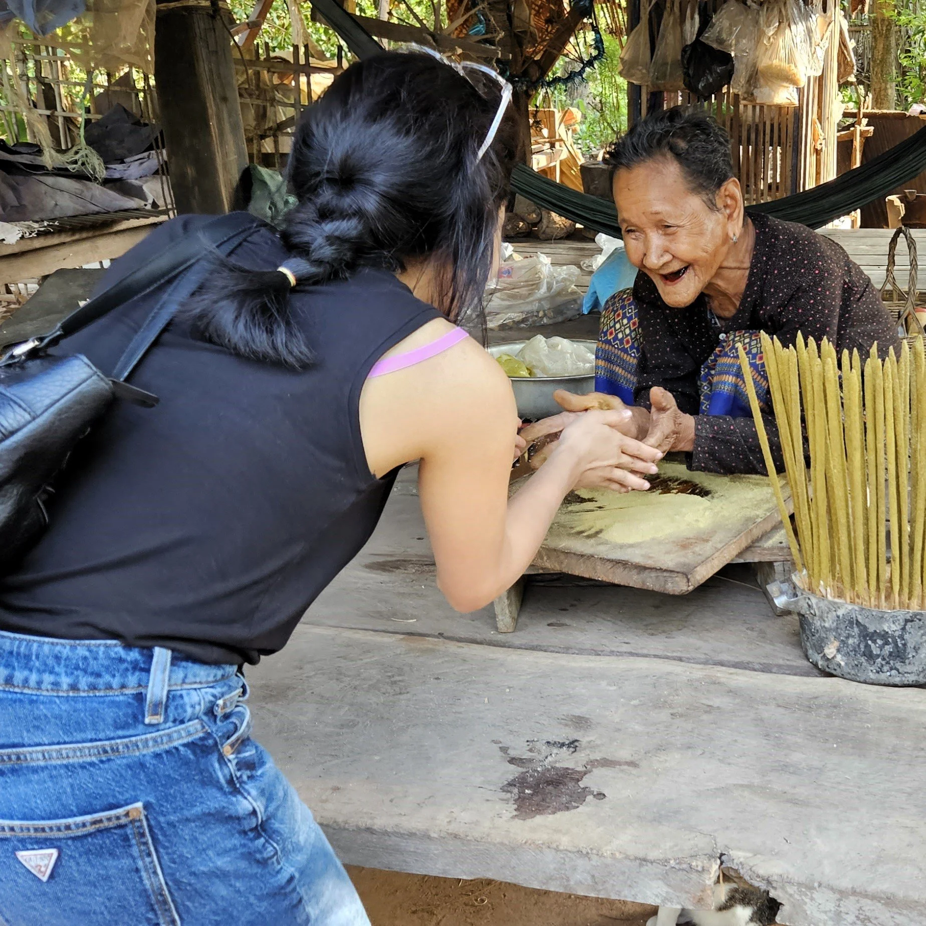 A young woman and an elderly woman sharing a joyful moment, holding hands across a wooden table, in a rustic outdoor setting with trees and wooden structures.