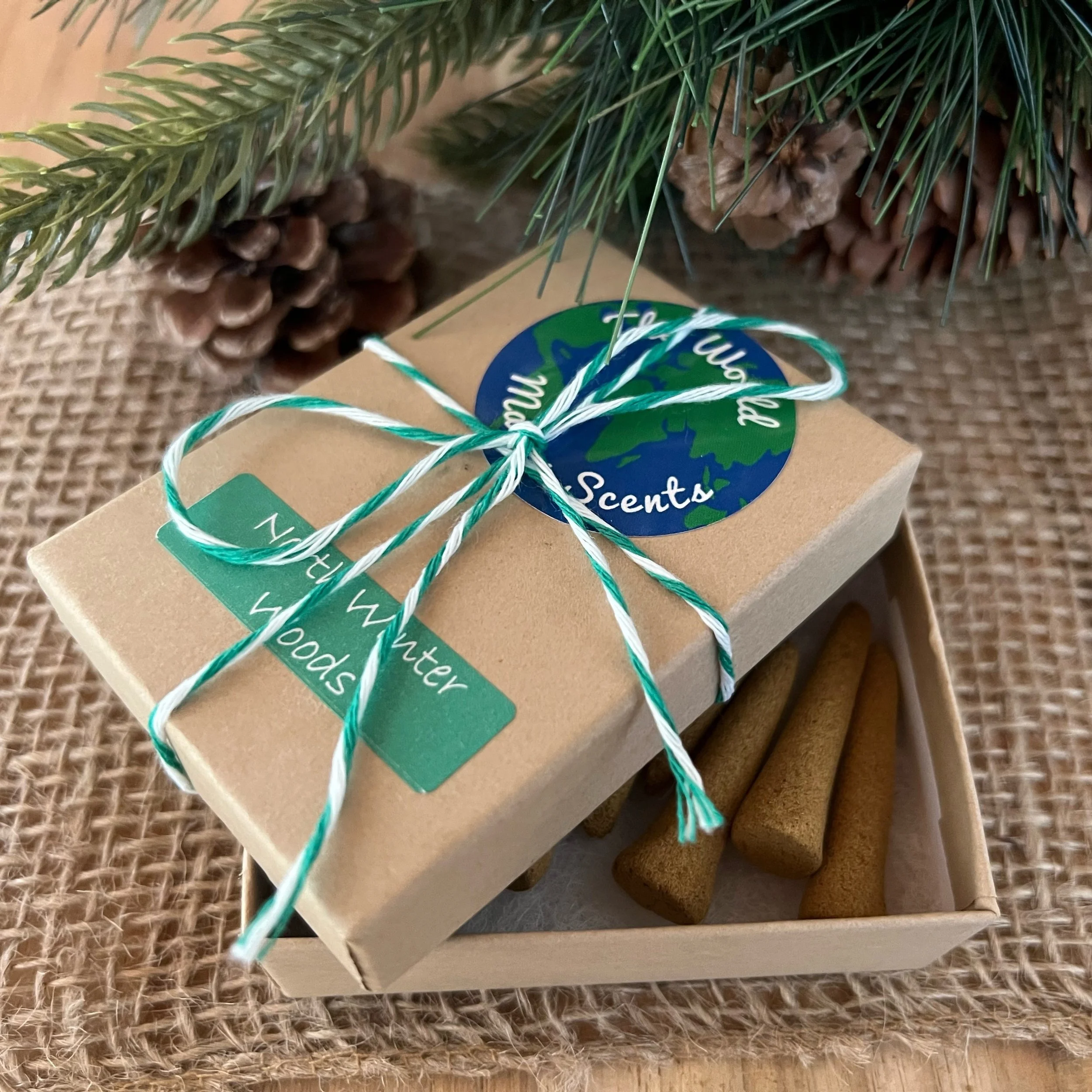 A collection of Christmas decorations including pine branches, pinecones, and a box of scented products tied with a green and white string, placed on a woven surface.