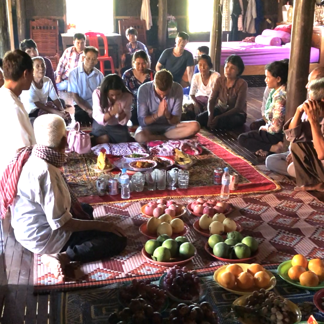 Group of people sitting on the floor in a circle during a religious or cultural ceremony, with offerings of fruits and food in front of them inside a traditional wooden house.