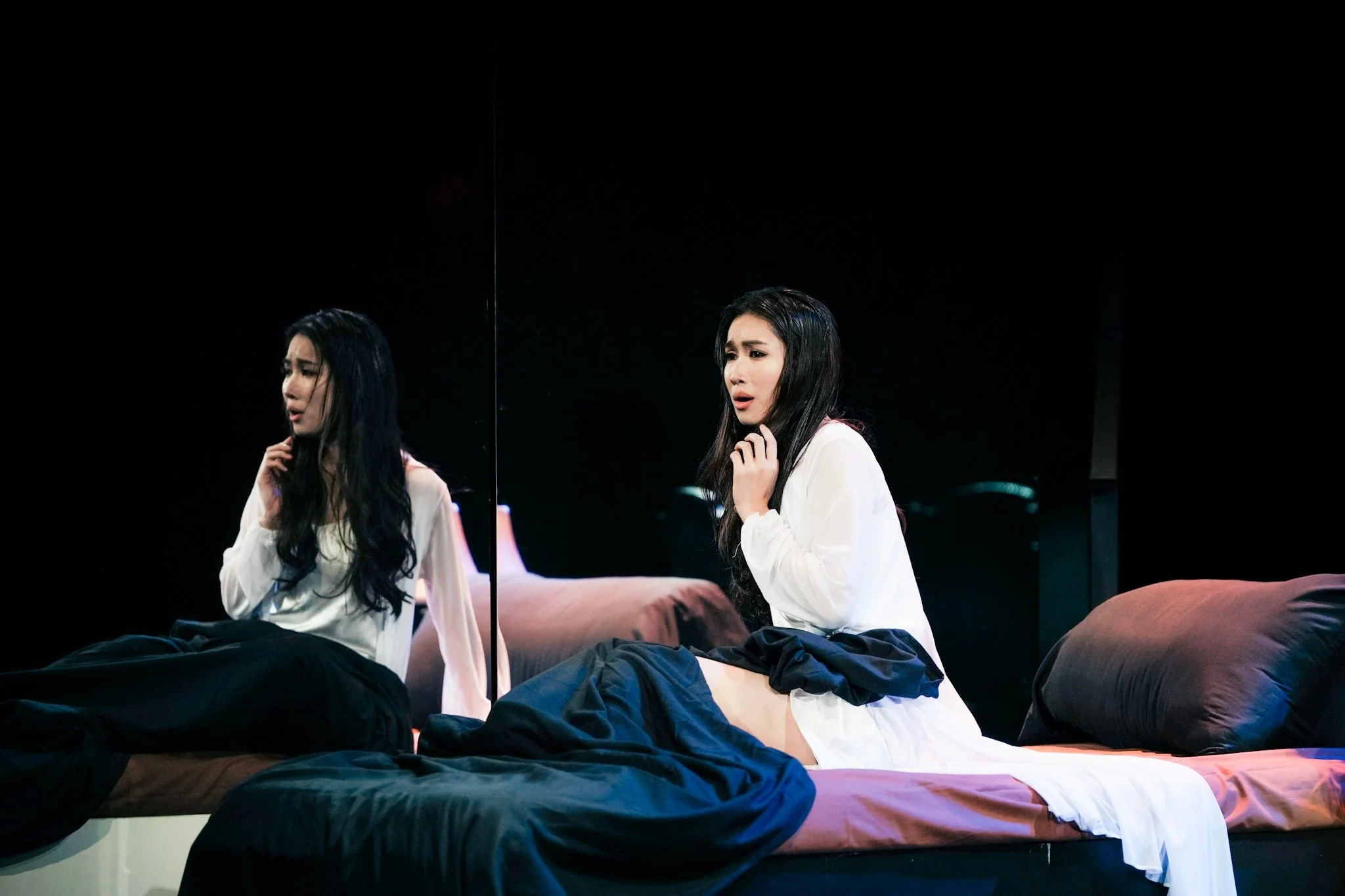 Two women with long dark hair sitting on beds with black bedding in a dimly lit room, looking distressed, with their hands near their faces.