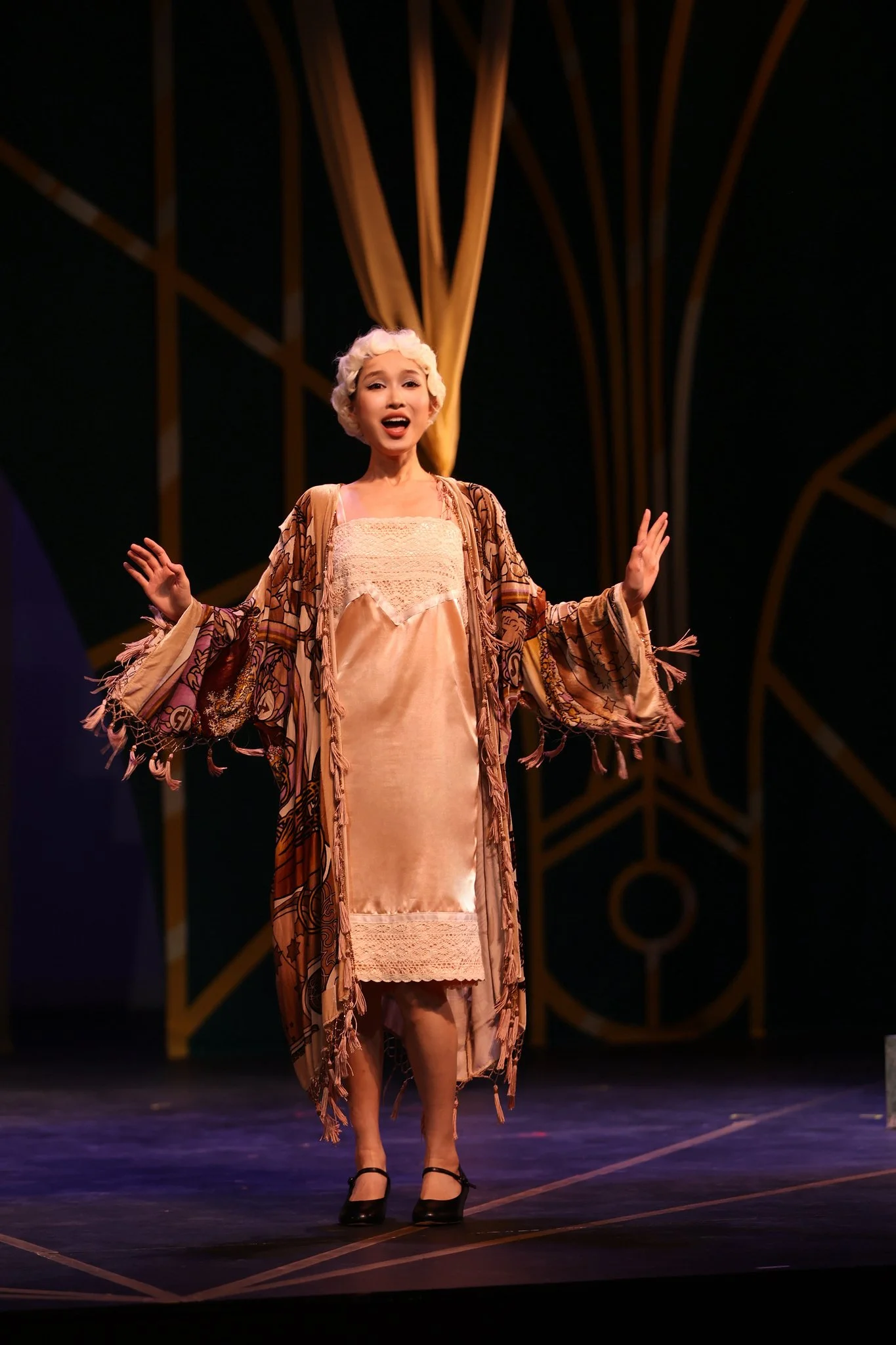 A woman on stage singing, wearing a peach-colored dress with lace detail, a patterned shawl, and black heels, in front of a stage set with wooden decorative elements and a dark background.