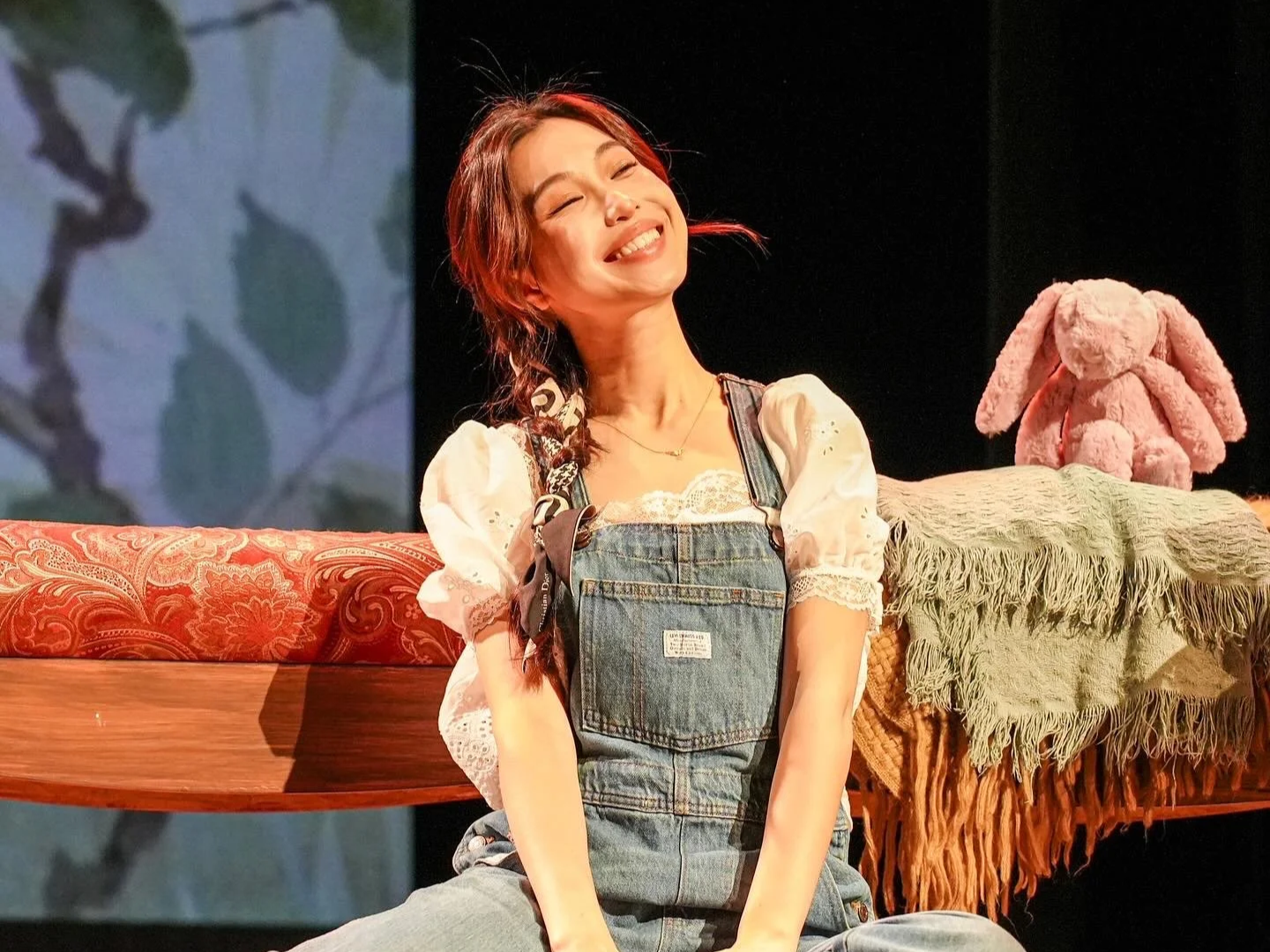 A woman smiling with closed eyes, sitting on a red-patterned sofa, wearing denim overalls over a lace-trimmed blouse, with a pink stuffed bunny and a table with a green fringe cloth behind her.