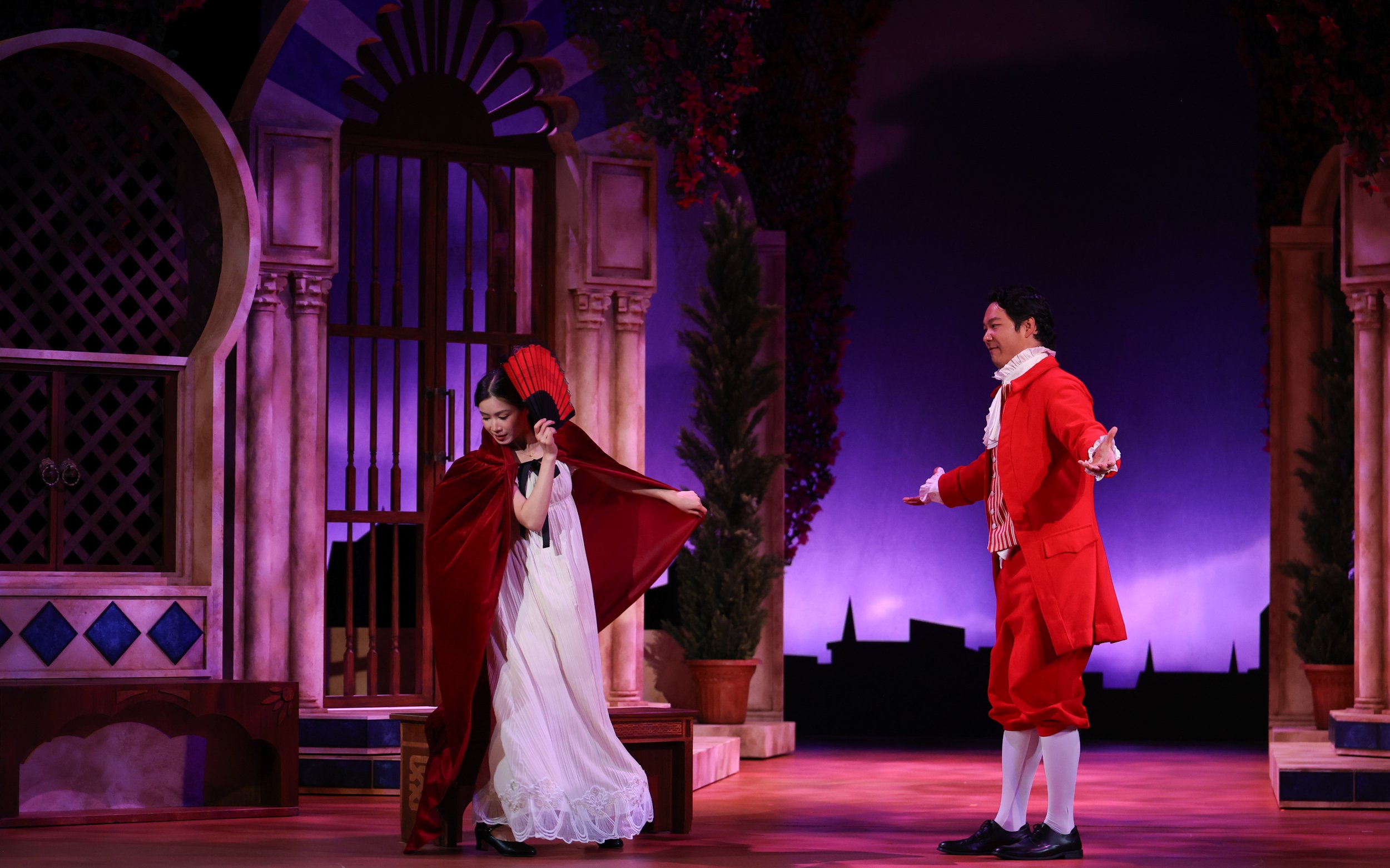 Two actors performing on stage in a theatrical play, with a woman dressed in a white dress and red cape holding a fan, and a man in a red suit with white ruffled shirt extending his hand, set against a backdrop of a garden with an ornate pink and pur