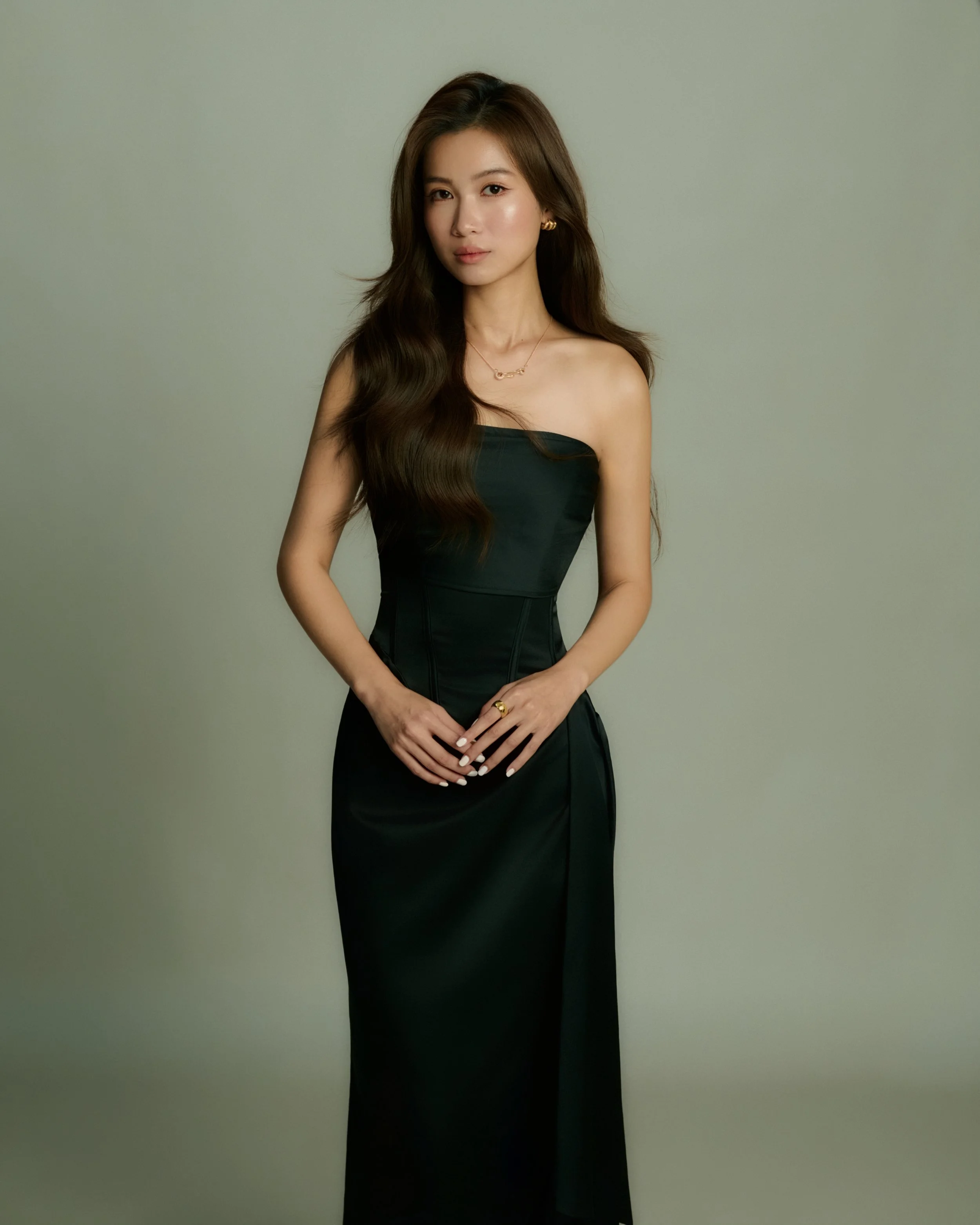 A woman with long, wavy brown hair wearing a strapless black dress, gold earrings, and jewelry stands against a plain light green background.