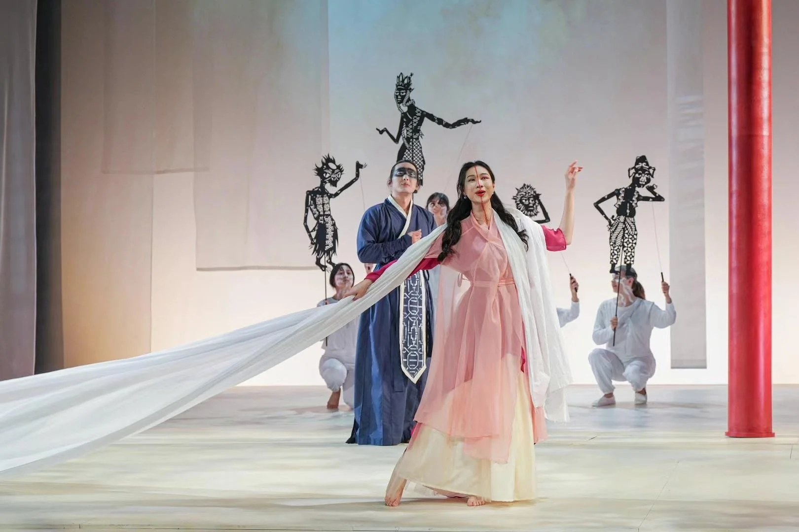 Performers in traditional costumes on stage with bamboo puppets in the background.